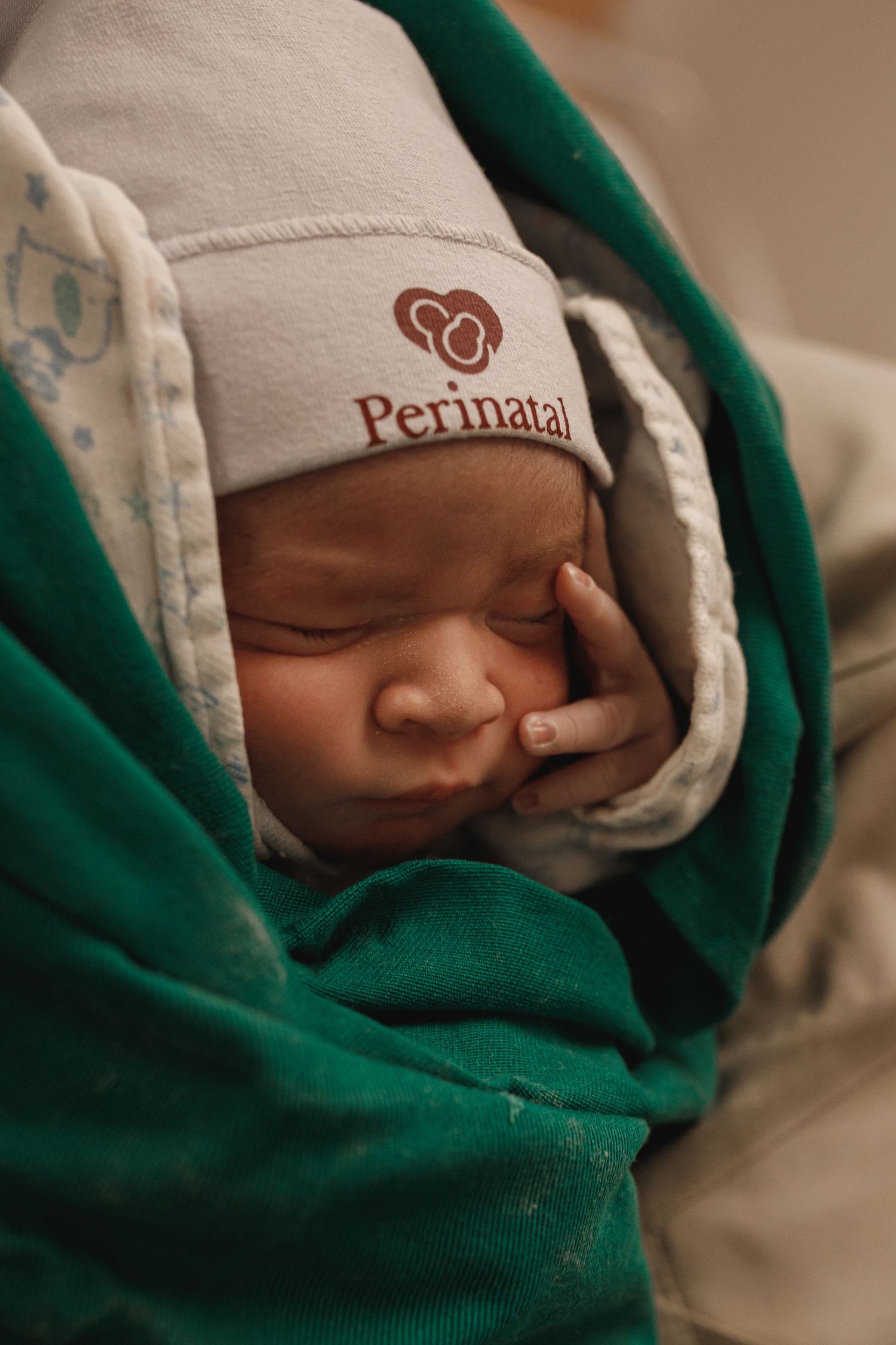 Close-up of a sleeping newborn wrapped in a green and white blanket with a hospital cap.