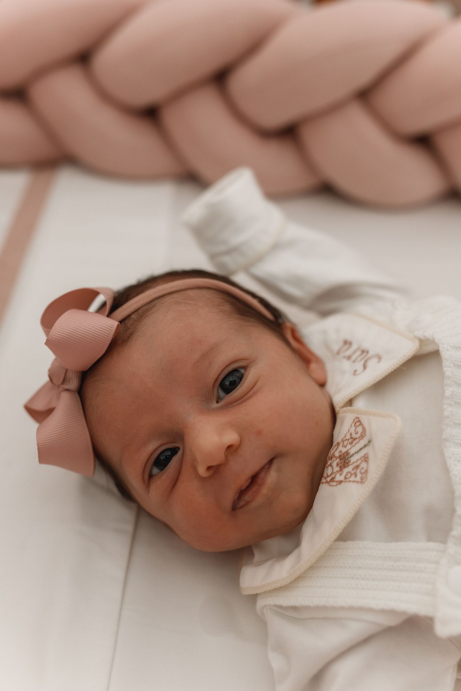 Newborn in white outfit lying against pink braided cushion backdrop.