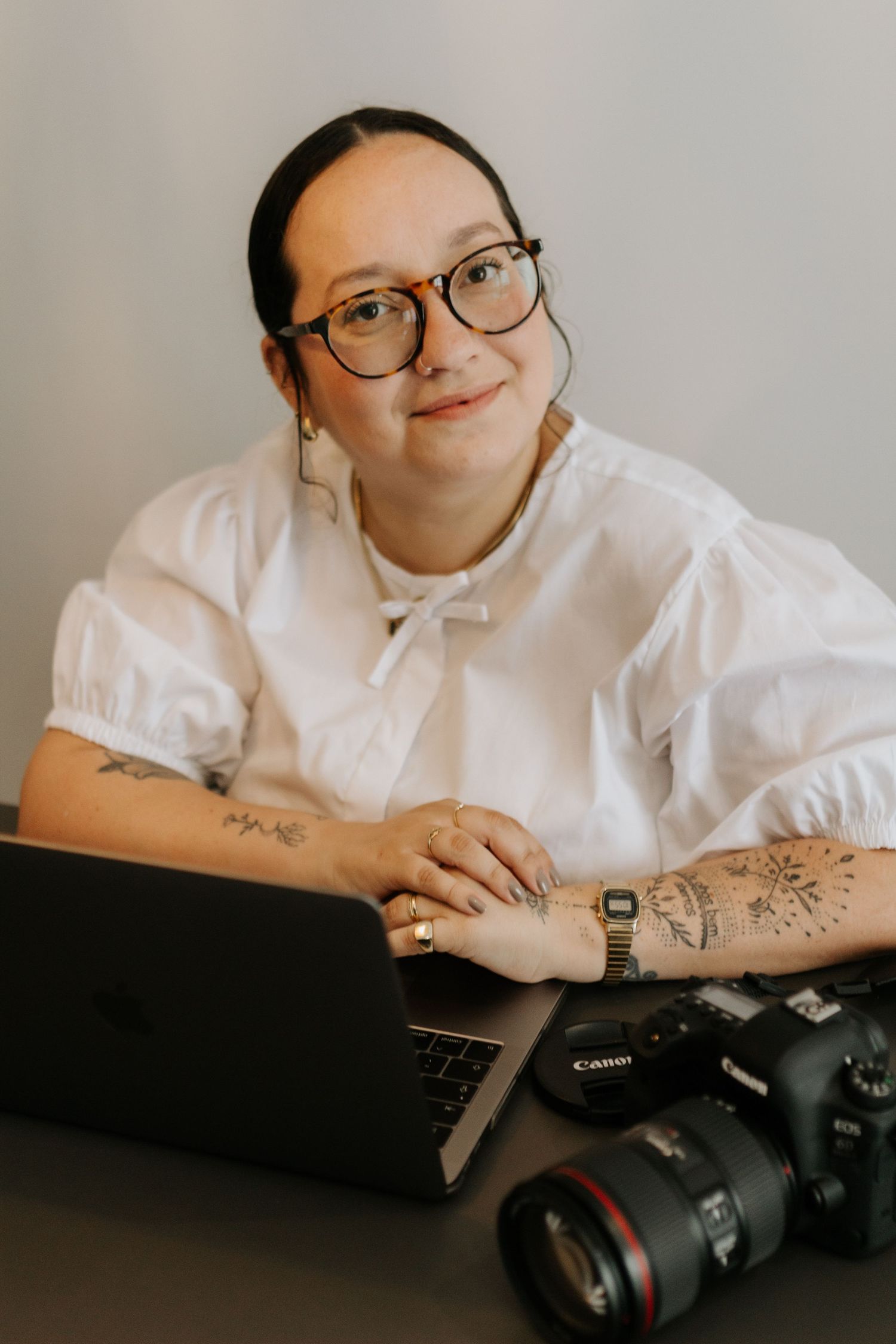 Photographer in white blouse with tattoos holding a Canon camera while working at a desk.