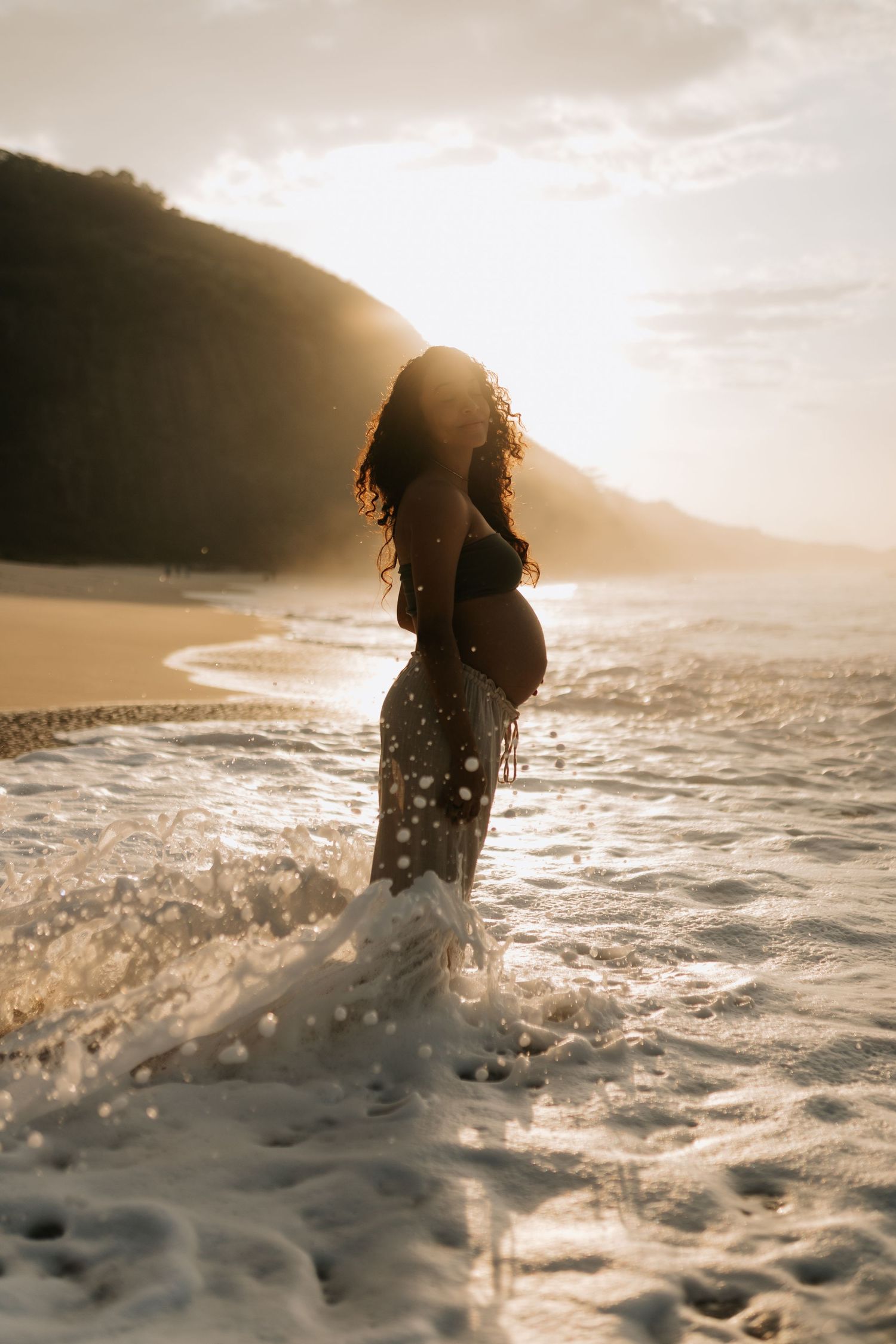 Silhouette of pregnant woman standing in ocean waves at sunset with mountains in background.