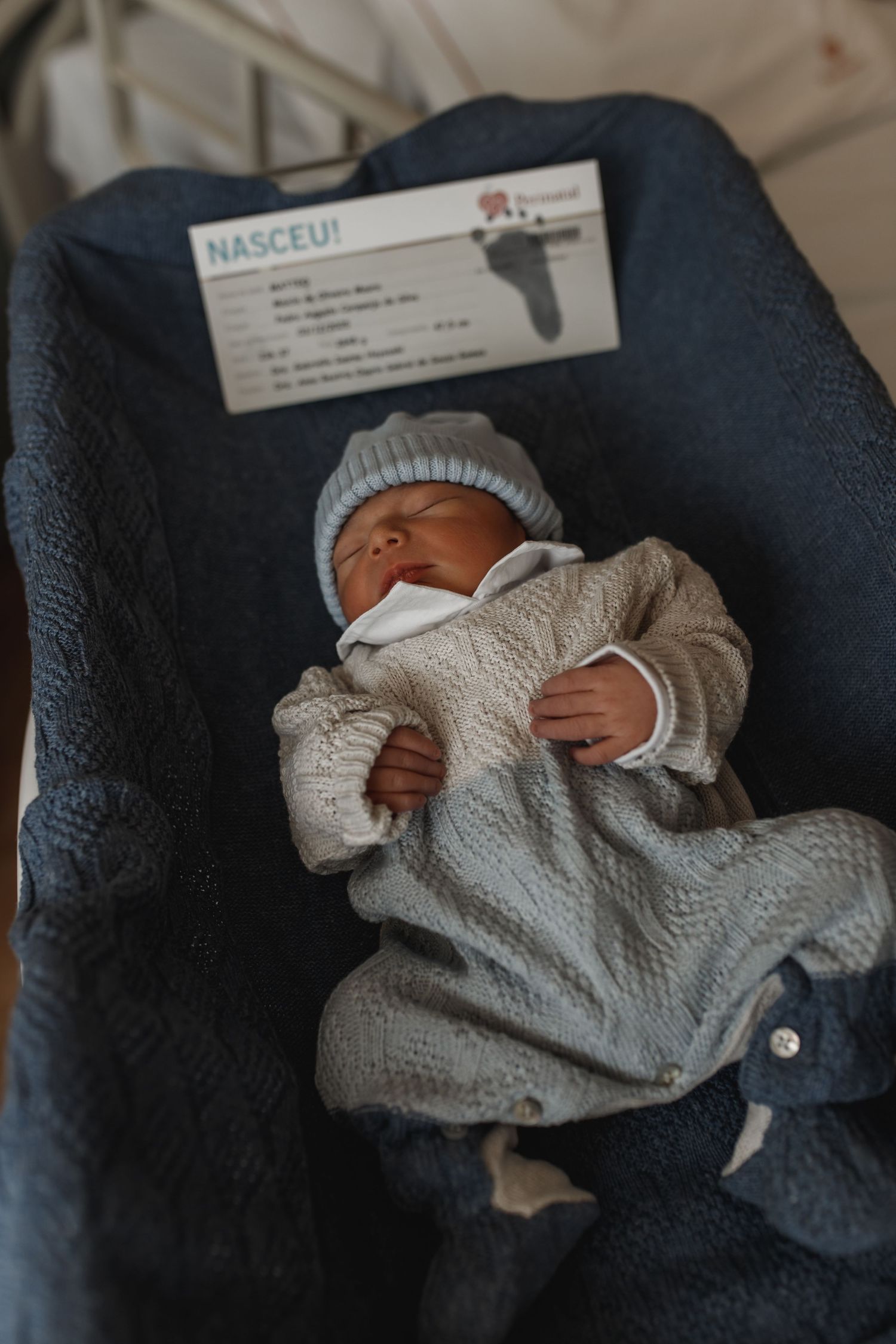 Newborn baby sleeping peacefully in a cozy gray knit sweater against dark fabric.