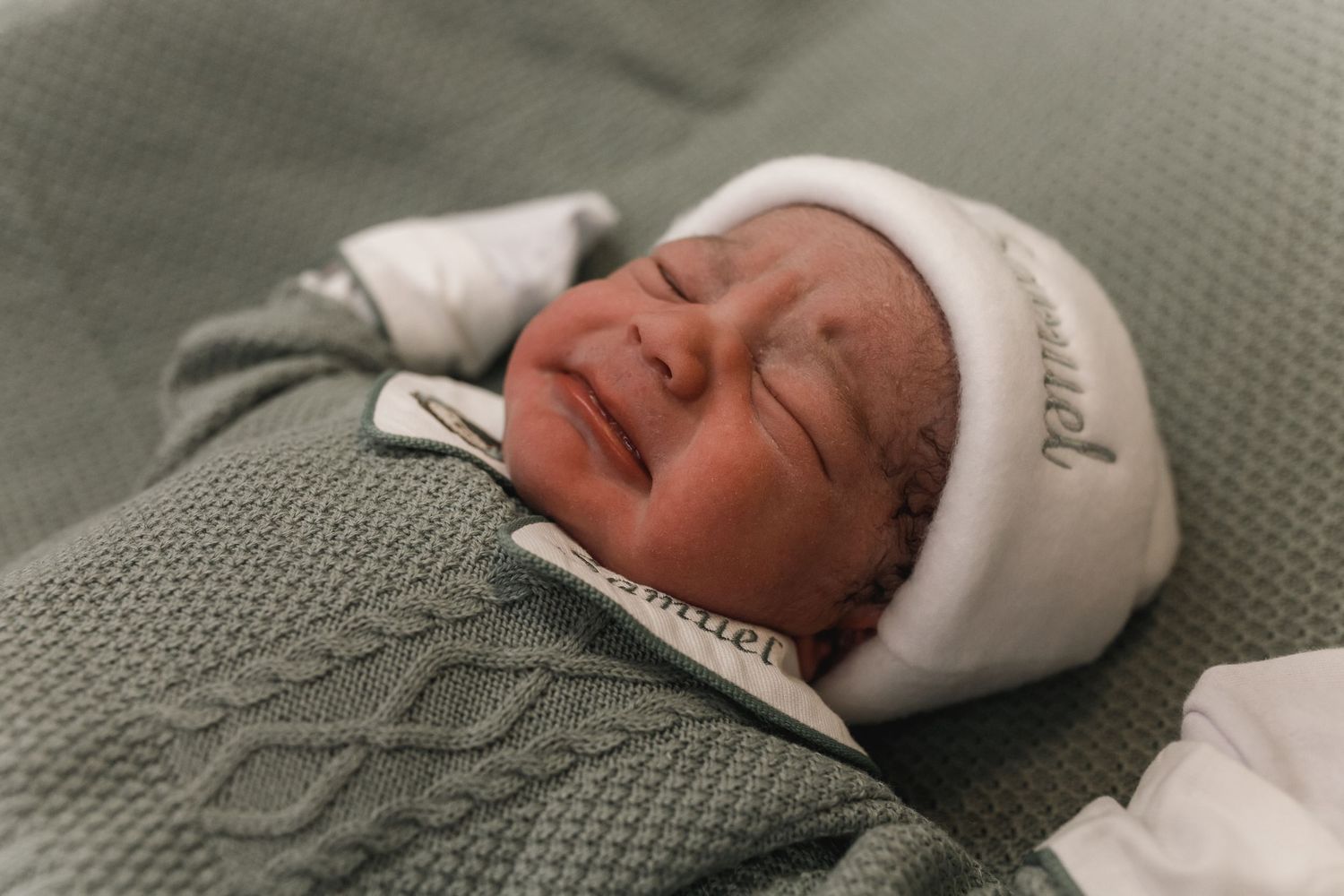 A newborn baby wearing a white cap rests on a textured gray blanket.