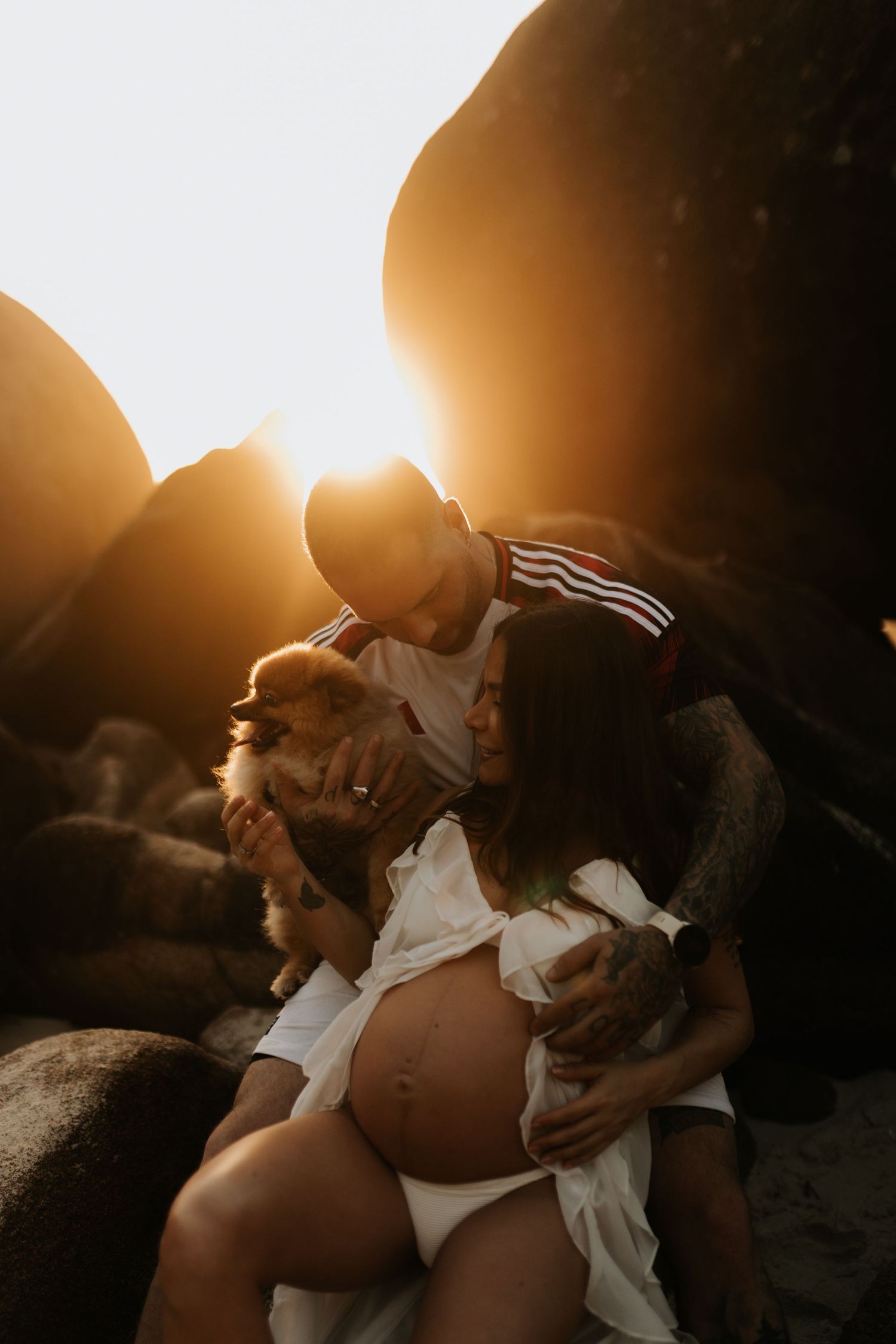 Silhouetted figures embrace against a bright sunset backdrop on a rocky beach.