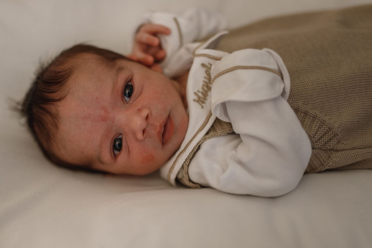 A baby in white clothing lies on beige bedding looking directly at the camera.