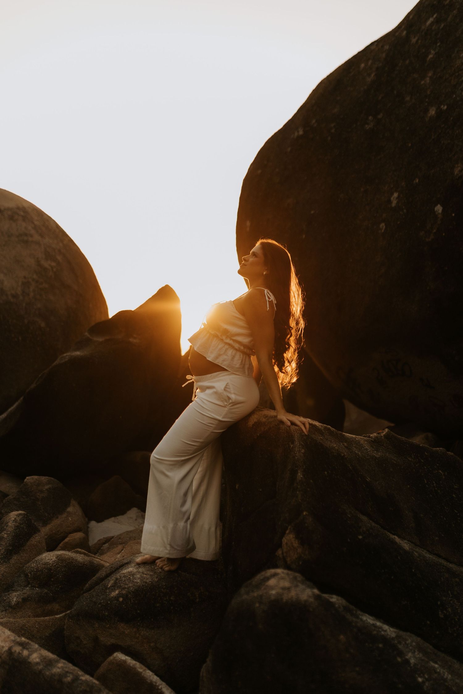 Silhouetted figure in white dress poses against rocky beach landscape at golden sunset.