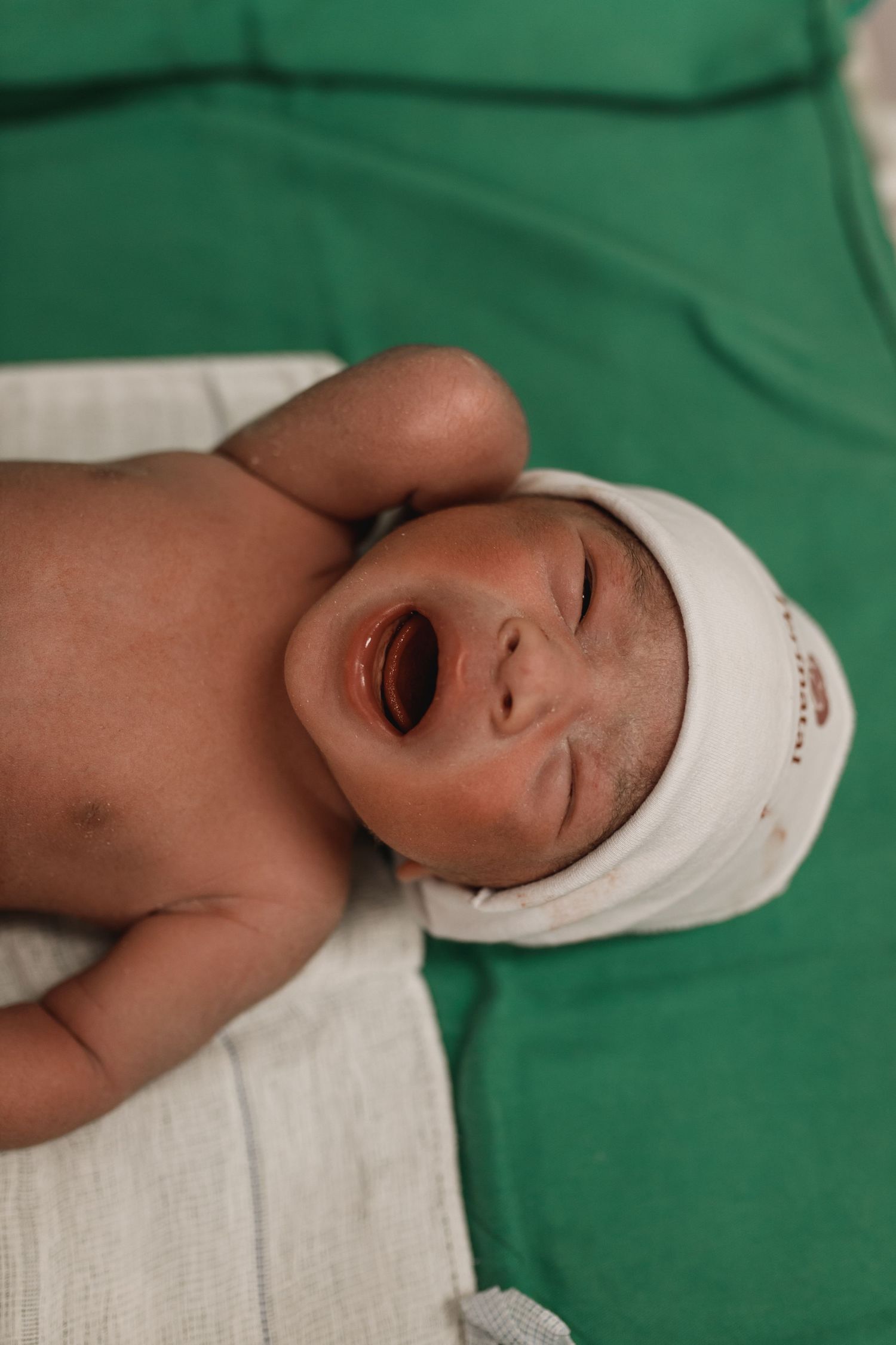 Newborn baby resting on green hospital bedding wearing a white cap.