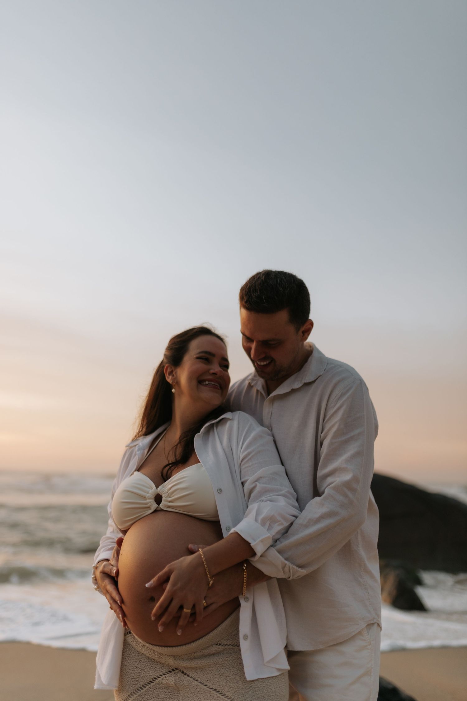 Intimate beach maternity photo of couple embracing at sunset with ocean waves behind them.