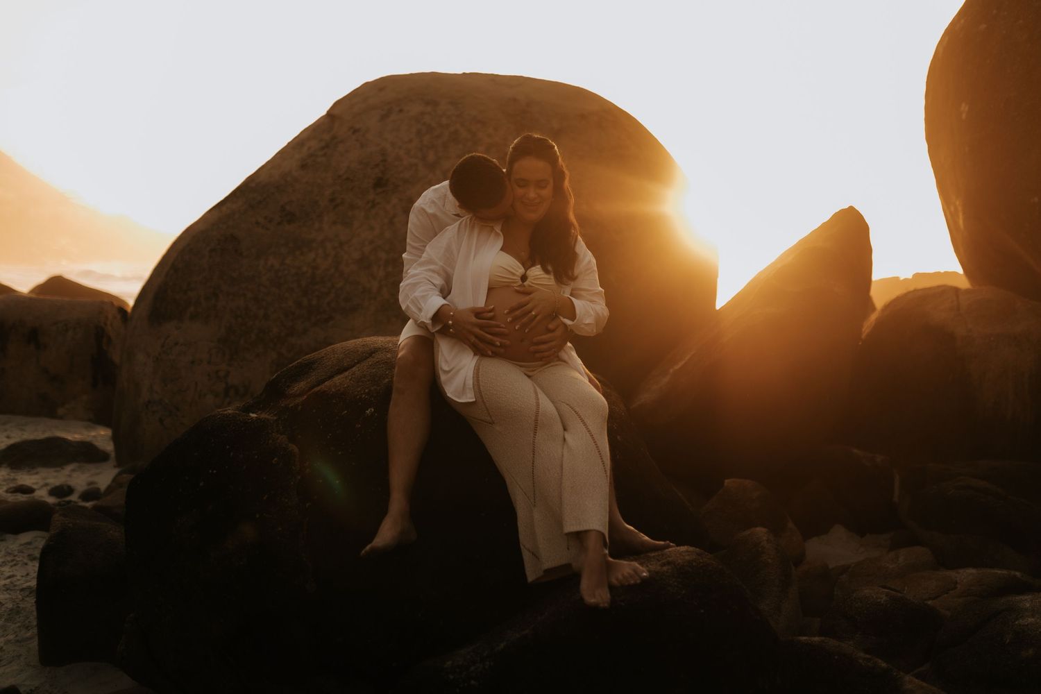 Romantic maternity photo of couple sitting on coastal rocks at golden hour.