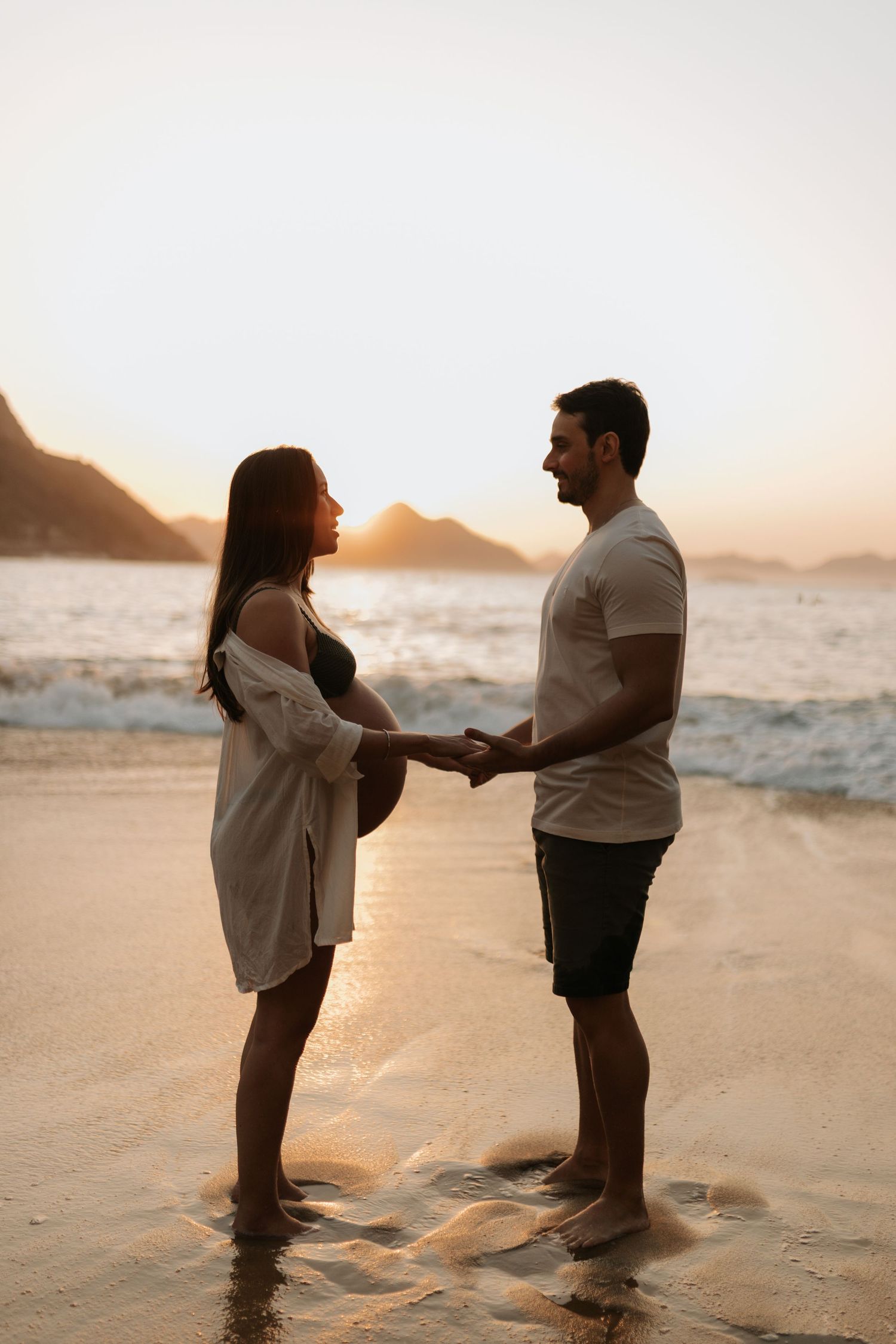 Two people stand facing each other on a beach at sunset with mountains in the distance.