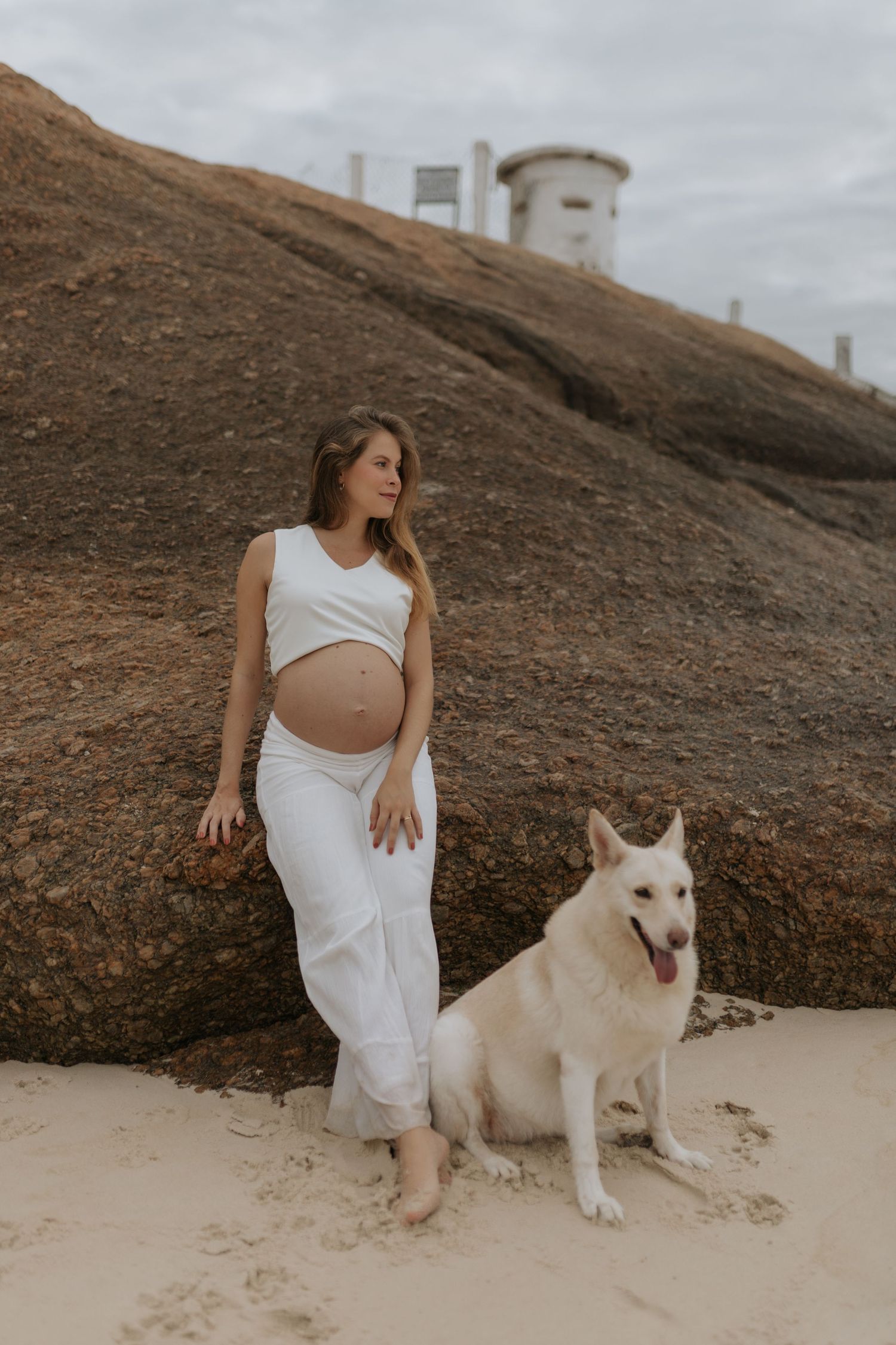 A person in white clothing poses with a white dog on a sandy beach near rocky cliffs.
