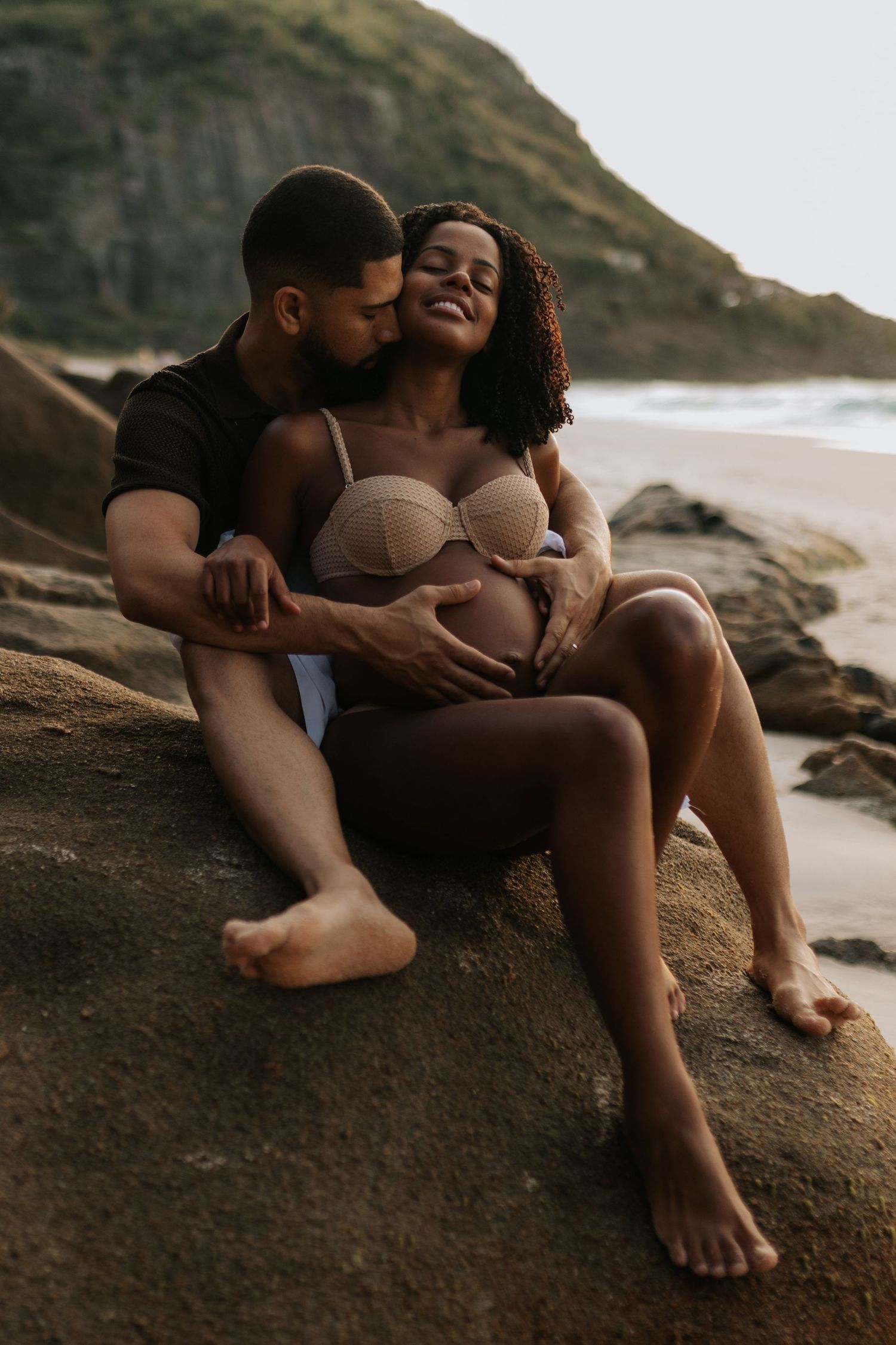 Couple shares tender moment on coastal rocks during sunset maternity session.