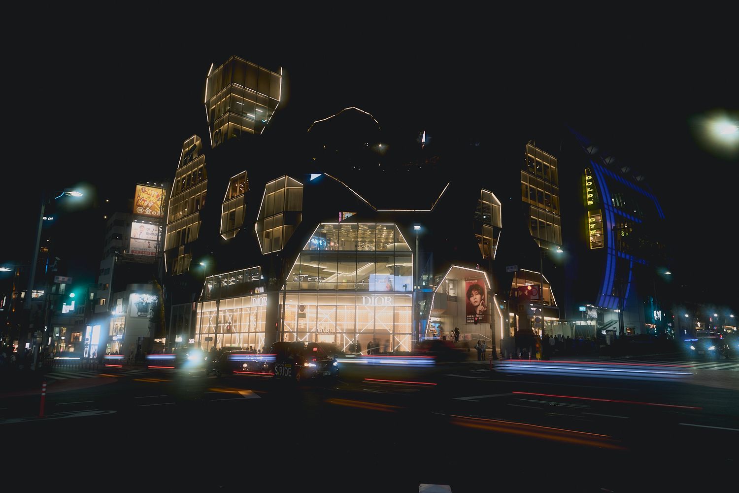 Illuminated modern shopping complex with glowing glass facades and light trails from passing traffic on a city street at night.