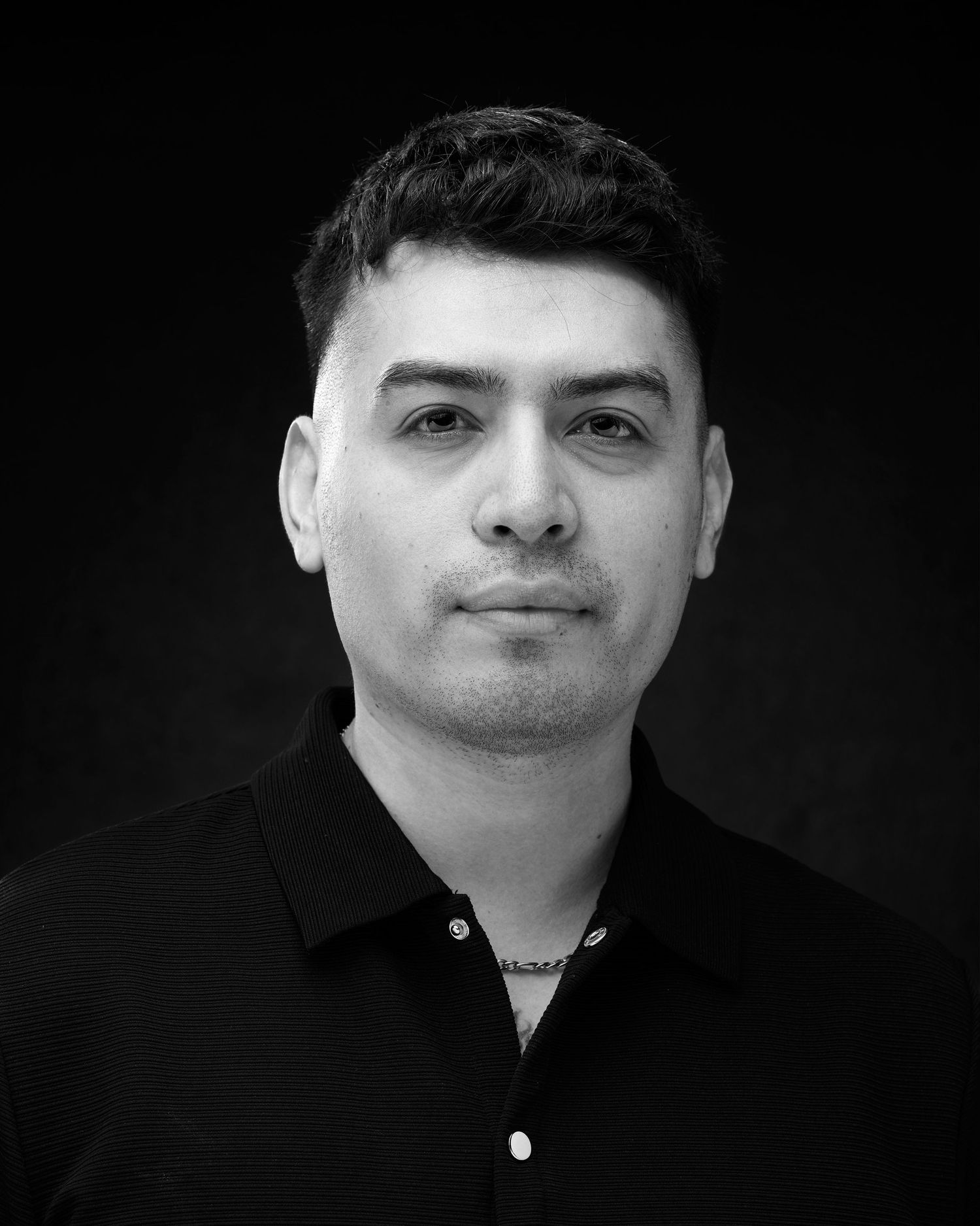 Black and white professional headshot of a young man in a black polo shirt against a dark background.