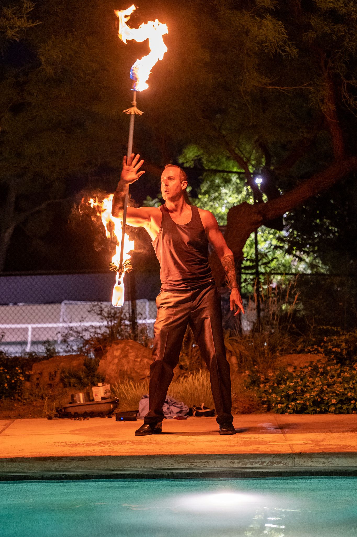 Fire performer spinning flaming torches near illuminated swimming pool at night.