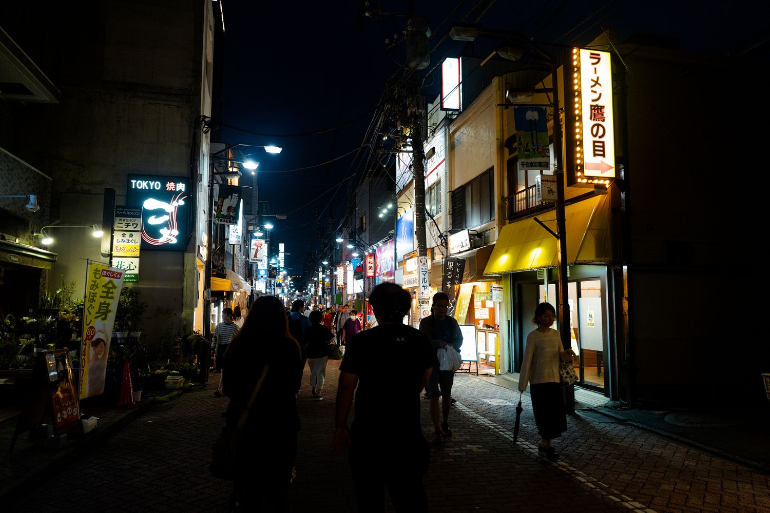 Nighttime street scene in Japan with illuminated shop signs and pedestrians.