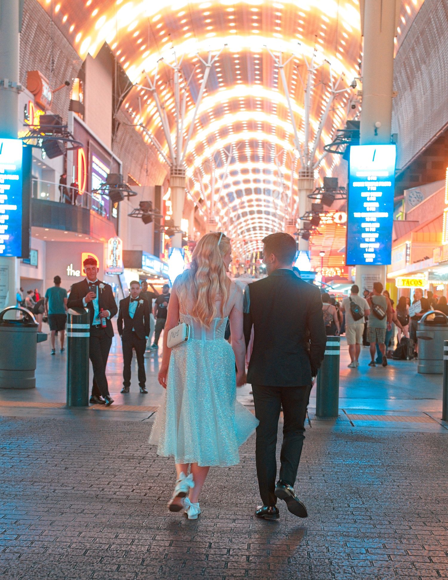 Brightly lit covered walkway with arched ceiling lights and pedestrians walking on cobblestone path.