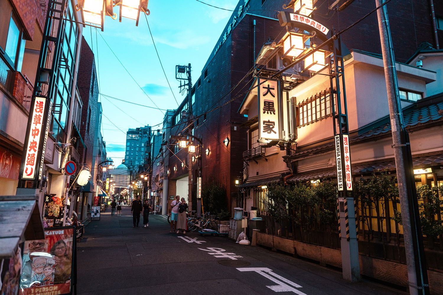 Japanese street scene at dusk with traditional lanterns and power lines against blue sky.