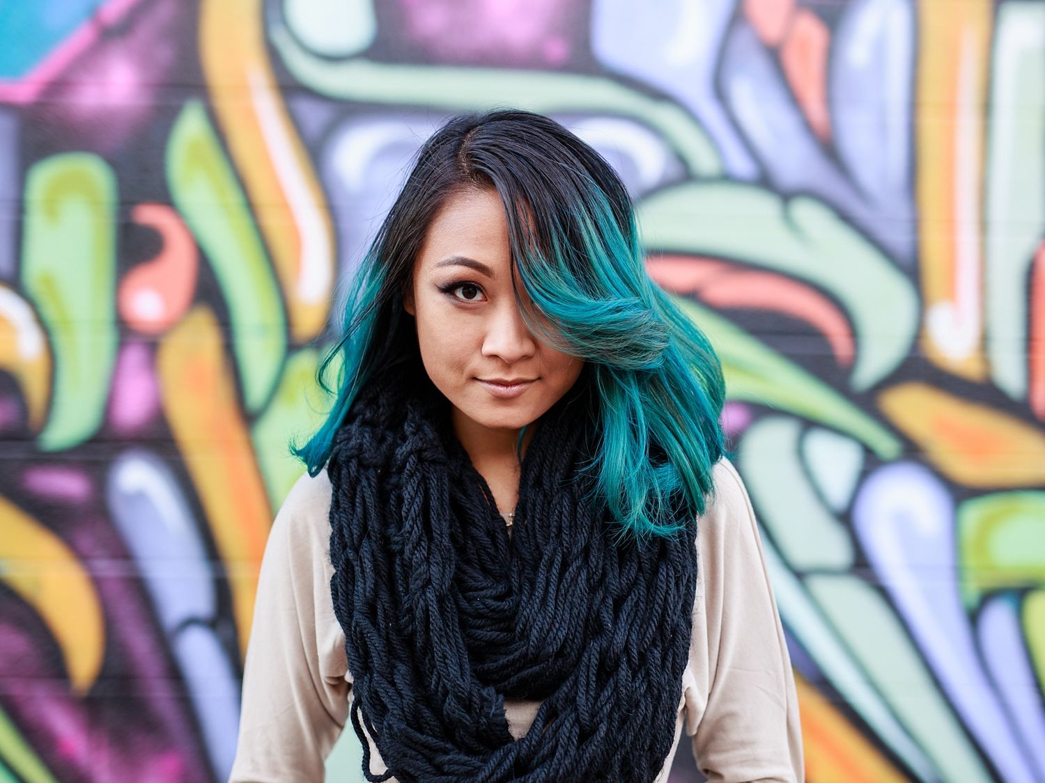 Person with vibrant teal hair standing in front of colorful graffiti wall with artistic street art designs.