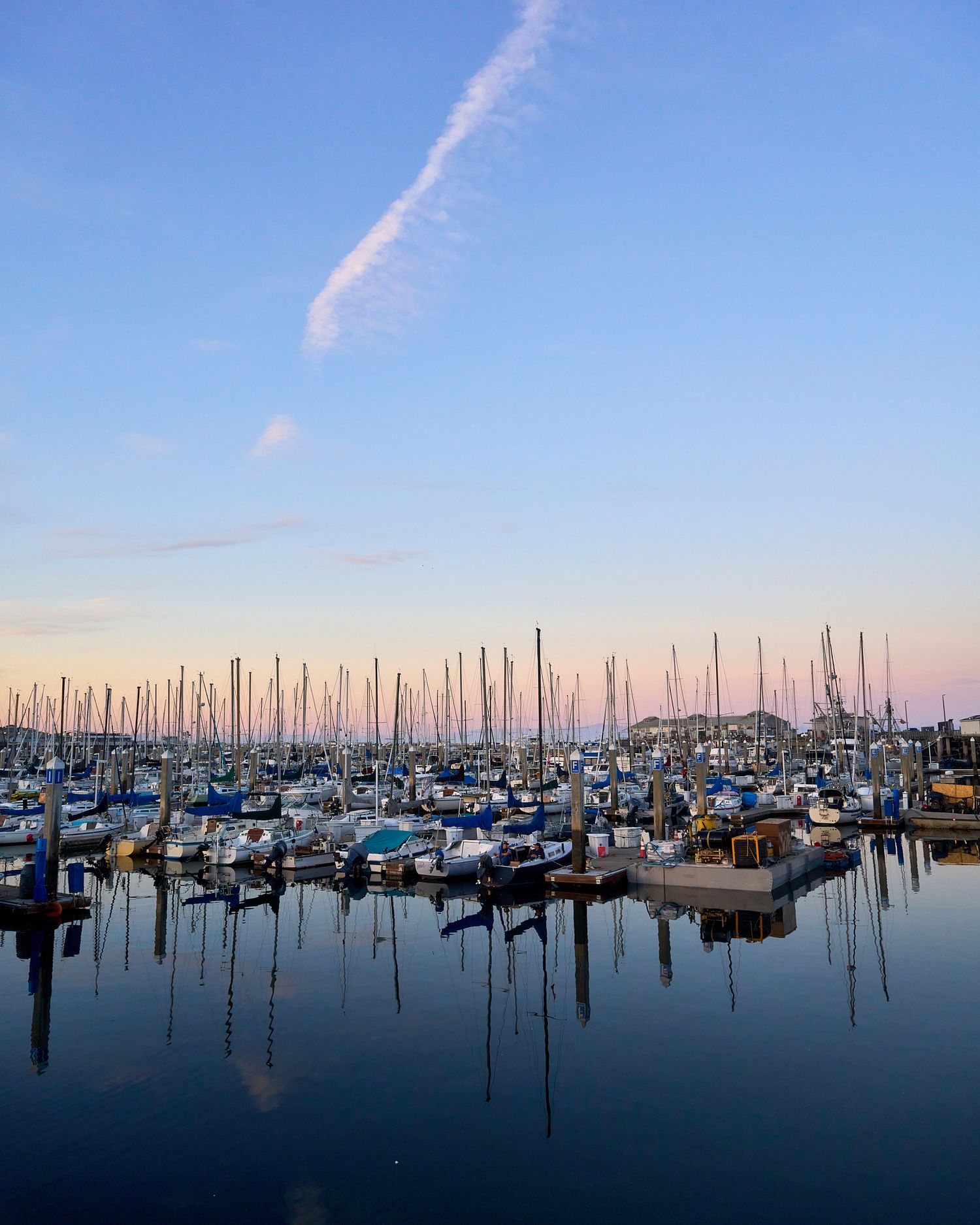 Marina filled with sailboats and yachts at sunset with masts reflecting in calm waters.
