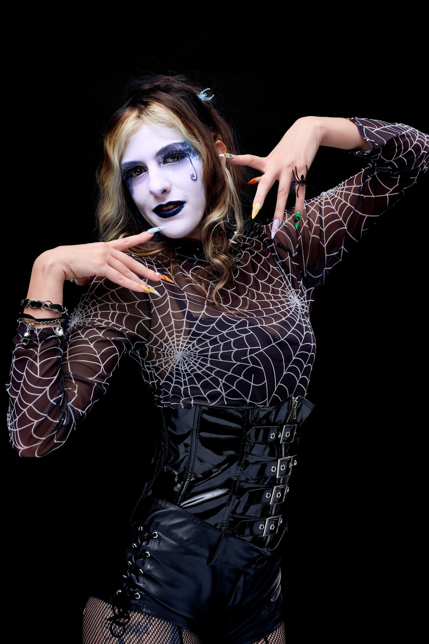 Woman in gothic Halloween costume with spiderweb bodysuit, black corset, white face paint, and long nails poses dramatically.