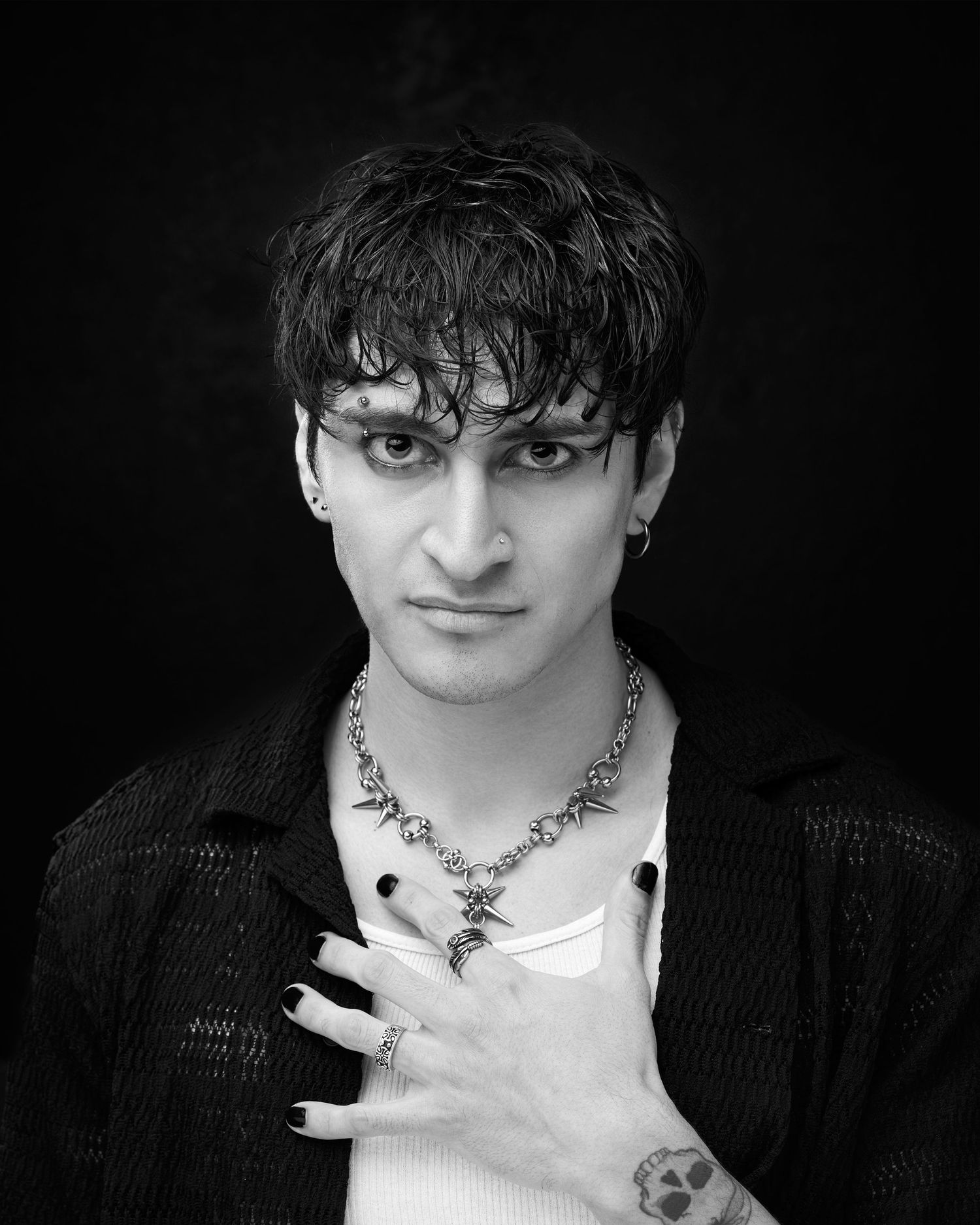Black and white portrait of a young man with dark hair, necklace, rings, and black nail polish posing dramatically.
