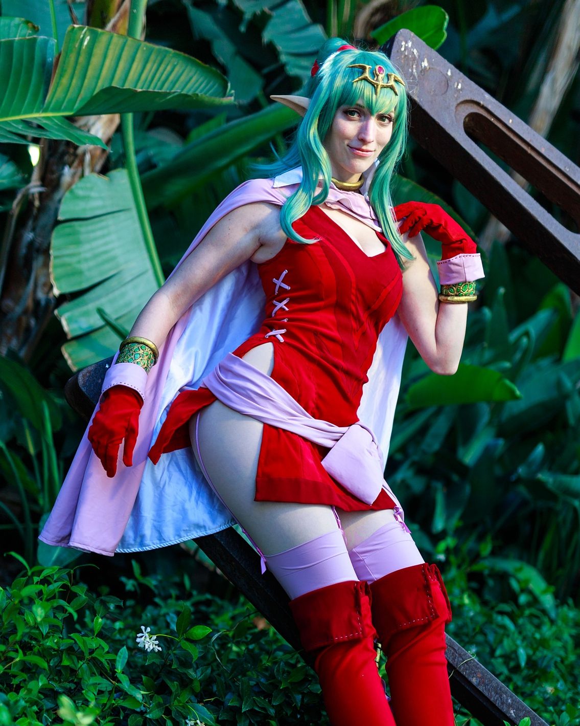 Cosplayer in red and white outfit posing in front of tropical foliage.