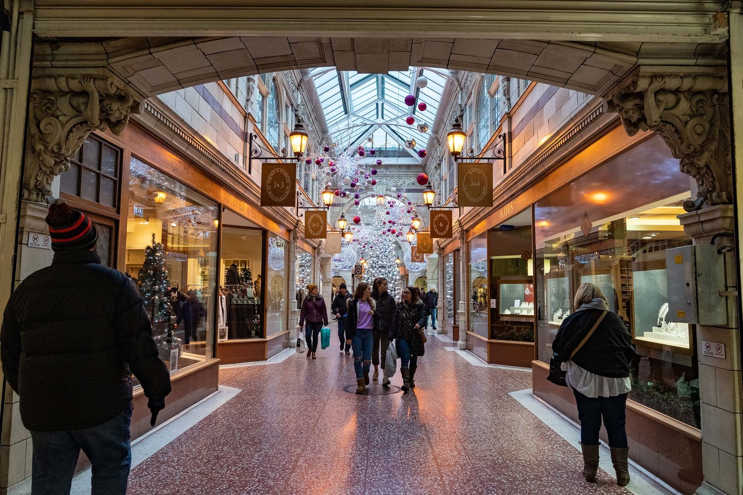 Indoor shopping arcade with glass ceiling and retail stores lining both sides of a long corridor.
