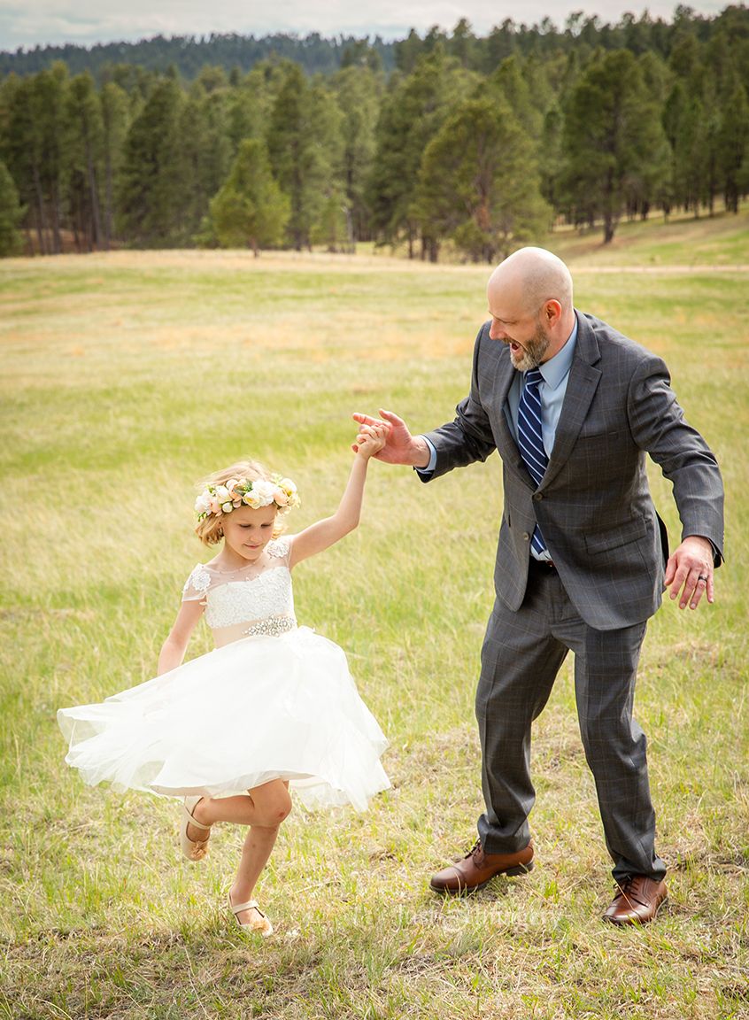 groom twirling and dancing with flower girl, pine trees and green field in the background