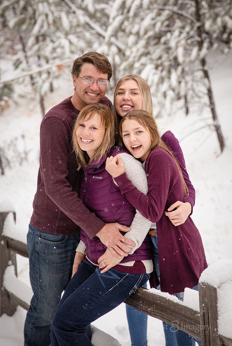family of four, with two daughters standing in the forest in the snow hugging each other