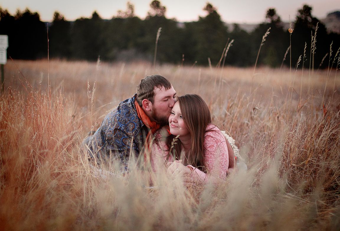 engaged couple lay down in a field at sunset, backlit, man kisses woman on the cheek
