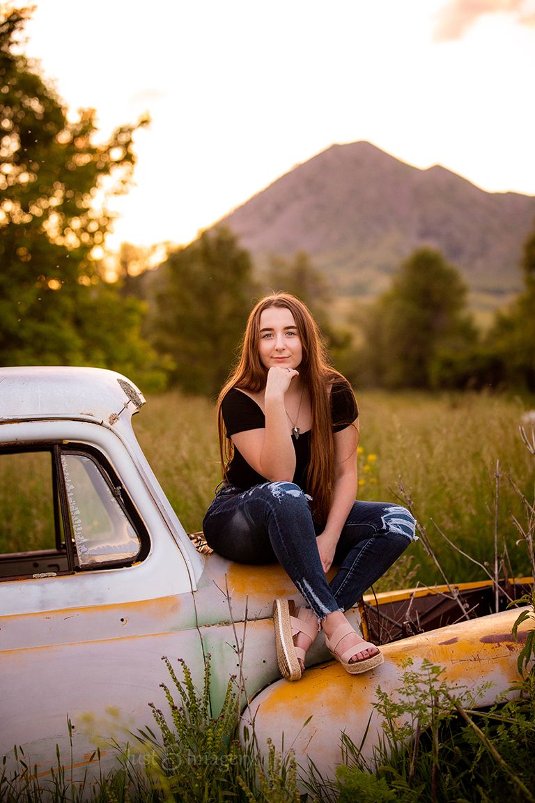 high school senior girl sitting on rusted vintage car with glowy trees and butte in the background