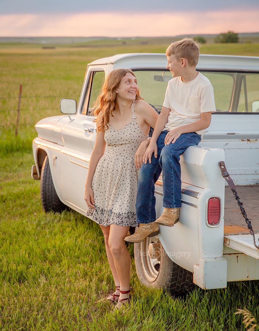 young brother and teenage sister smiling at each other while standing and sitting on the back of a classic white pickup truck