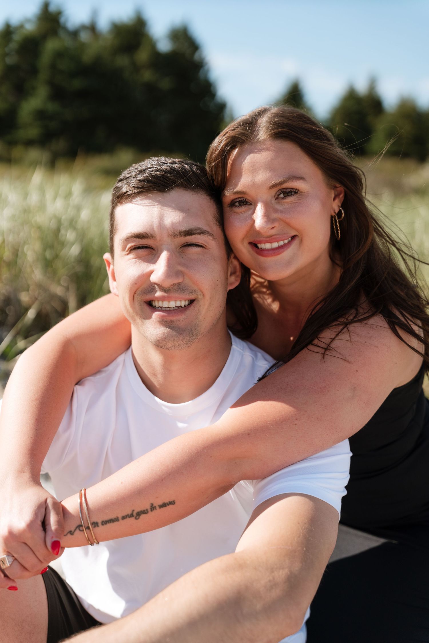Happy couple embracing outdoors, woman in black hugging smiling man in white tee, green trees background.