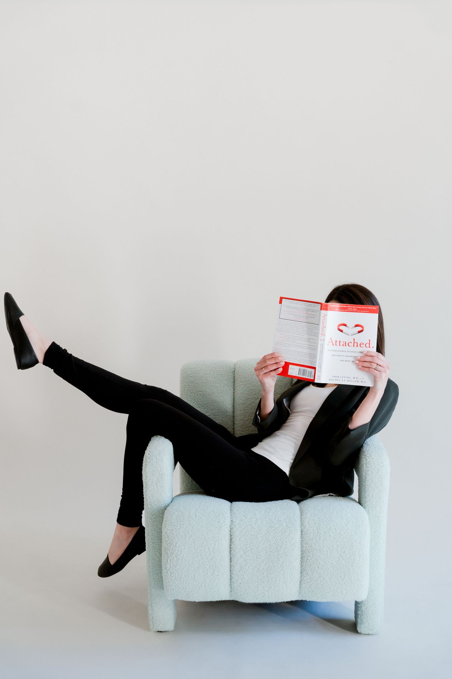 Woman in black suit sitting in a light blue armchair reading the book Attached, with one leg raised.