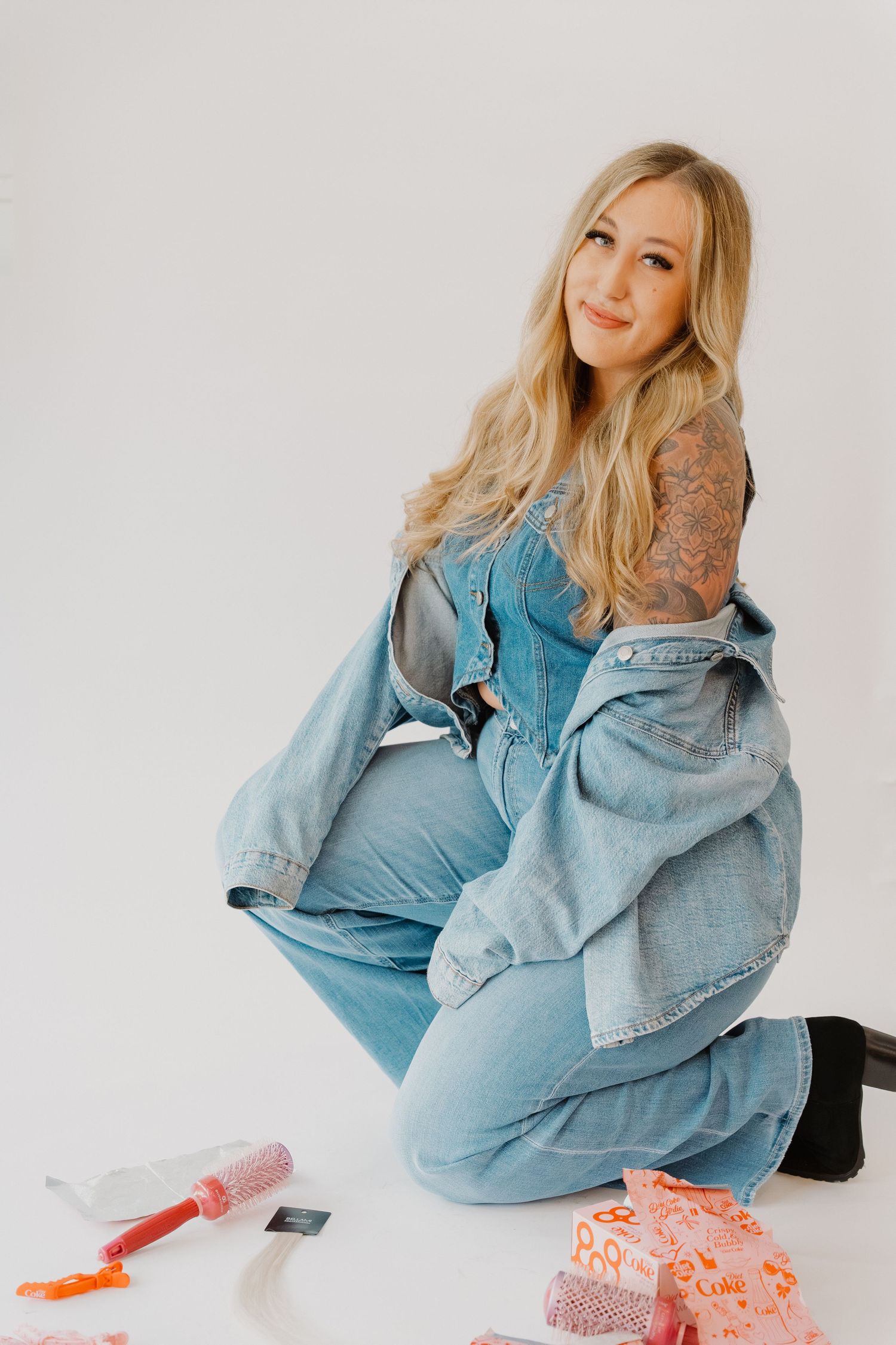 Pregnant woman with blonde hair and tattoos sits on white floor in denim overalls, surrounded by colorful gifts.