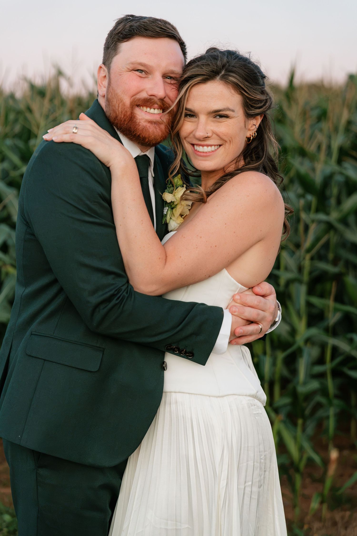 Smiling bride and groom embrace in front of a cornfield, groom in dark green suit with boutonniere.