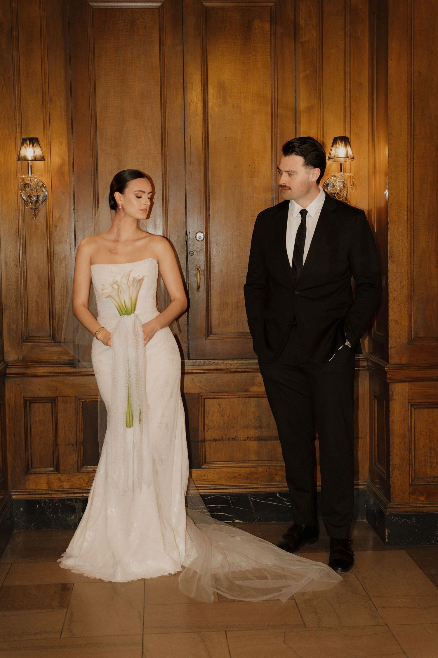 A couple in formal wedding attire poses playfully in a wood-paneled hallway for a series of candid photos.