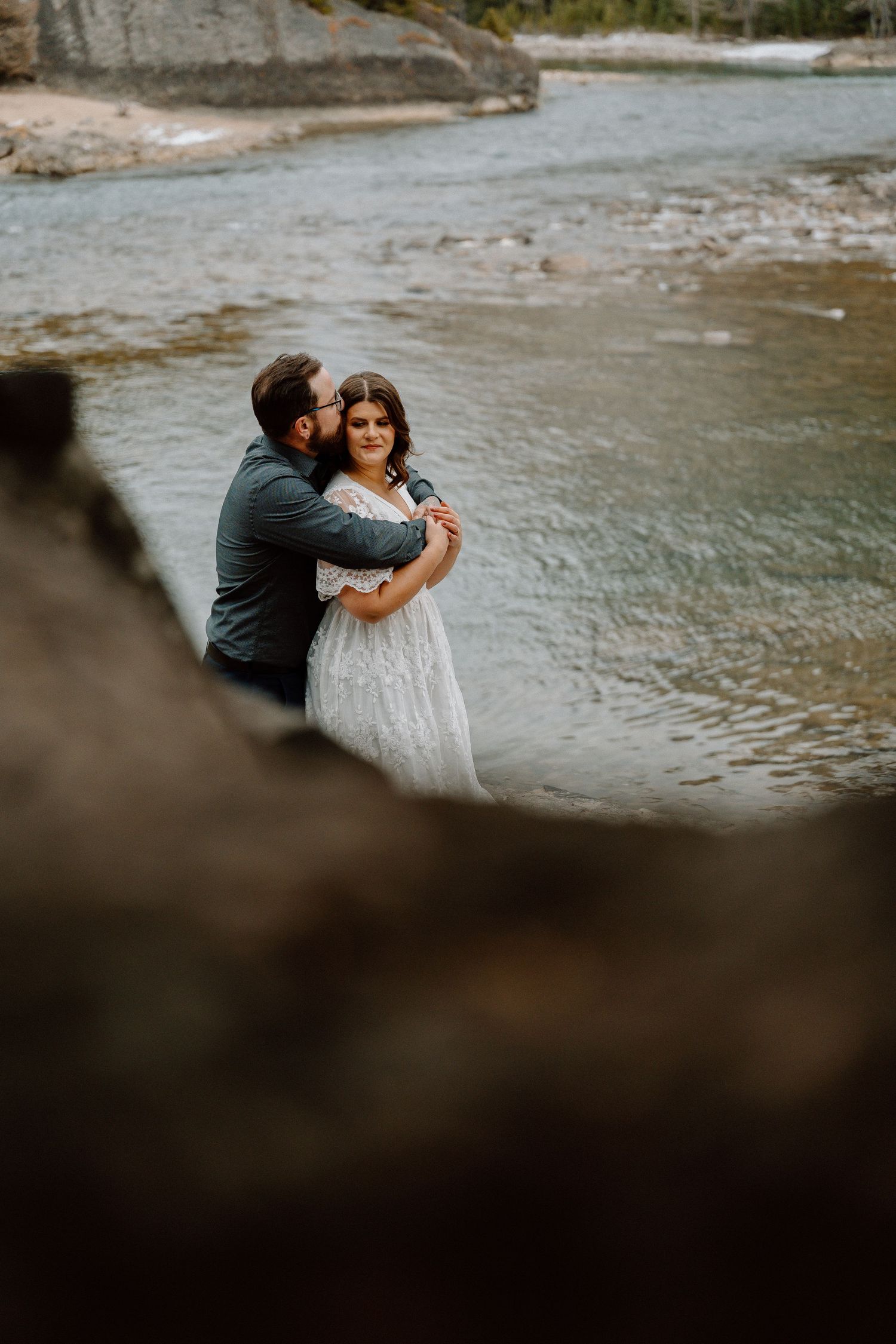 Couple embracing by a river with blurred foreground elements creating a romantic atmosphere.