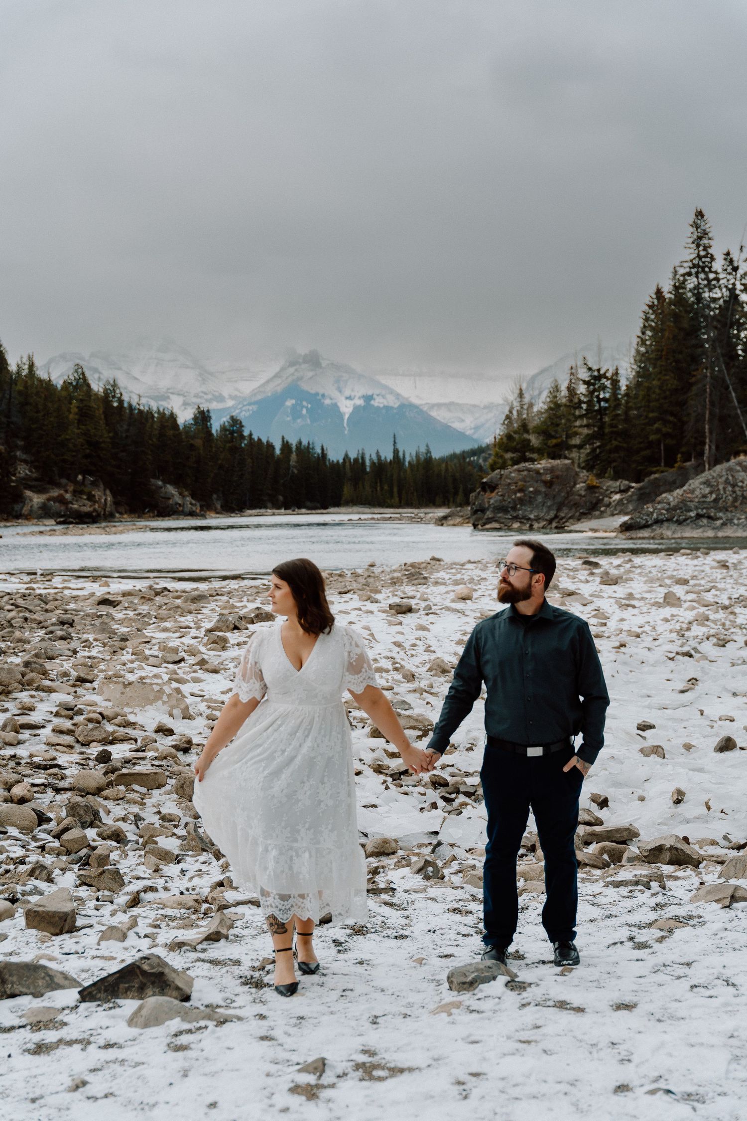 Wedding couple walking hand in hand across a snowy rocky shoreline with mountains in the distance.
