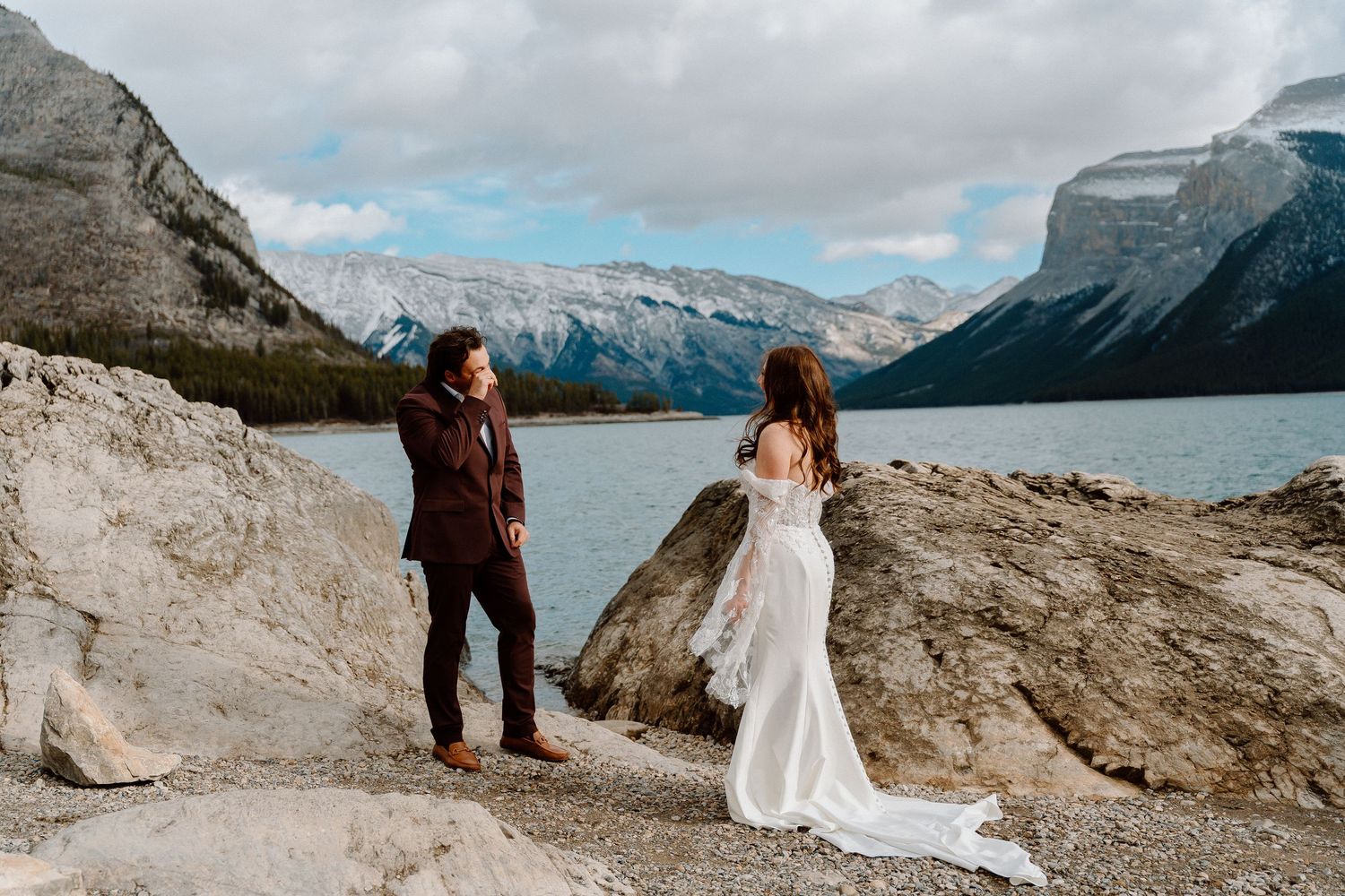 Couple in wedding attire poses along scenic mountain lake with dramatic cloudy skies and rocky shoreline.