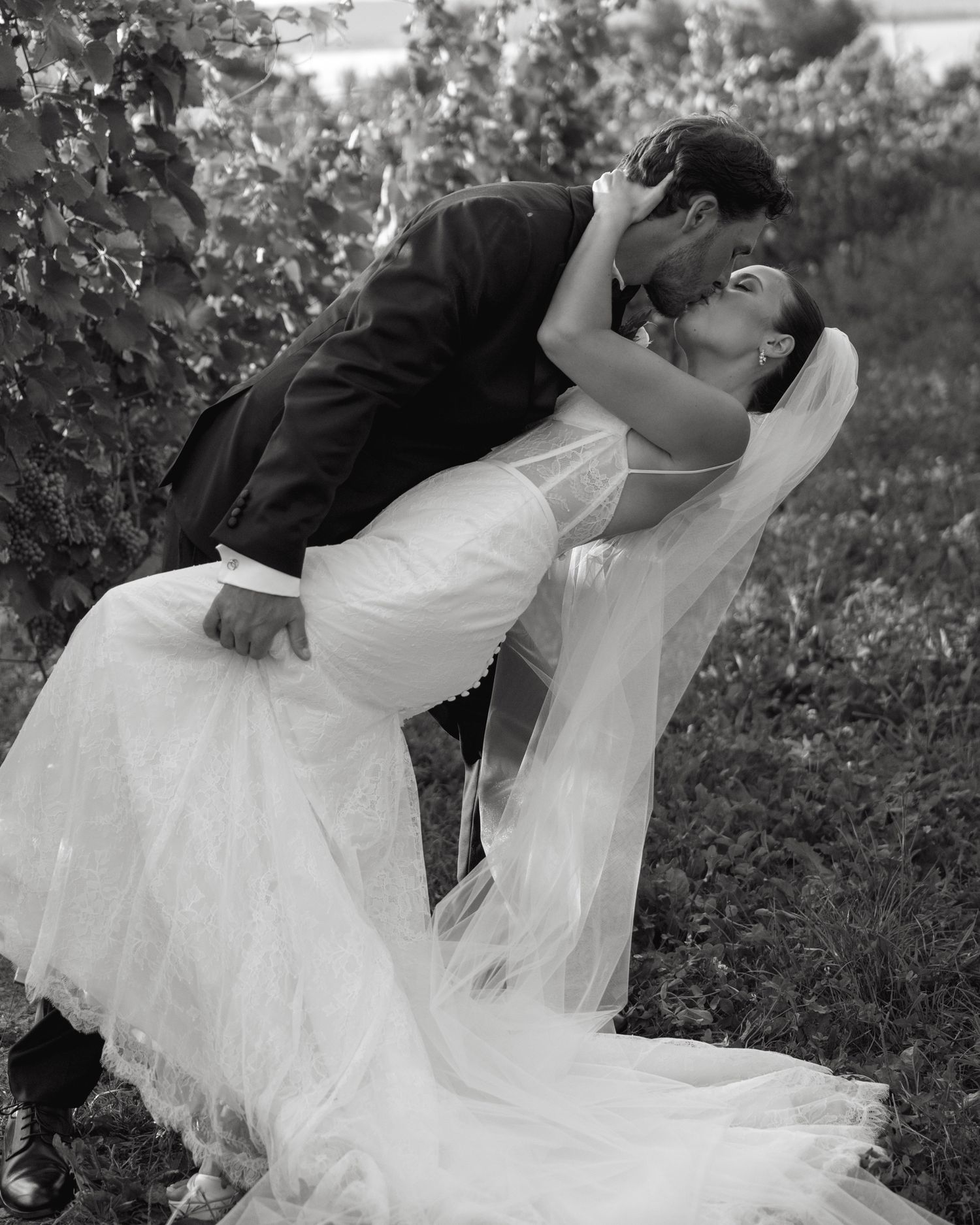 Black and white photo of a bride and groom sharing a romantic dip kiss in a lush vineyard on their wedding day.