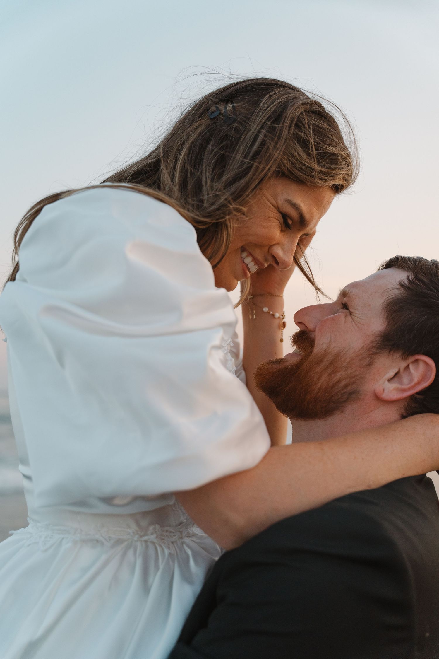 A joyful couple sharing a romantic moment outdoors, woman in white dress laughing as man gazes up at her lovingly.