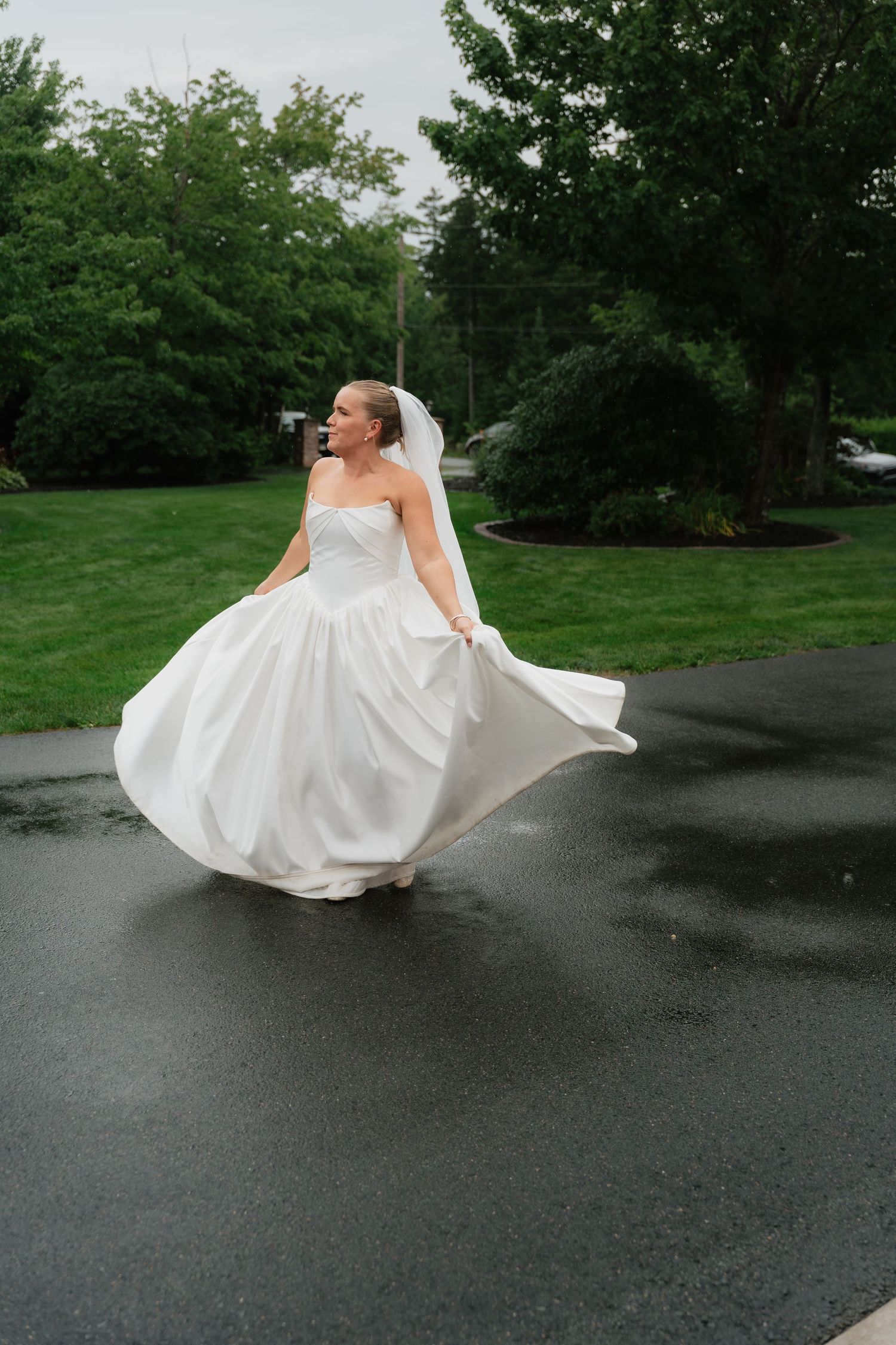 Bride in strapless white gown and veil twirling on wet pavement surrounded by lush green trees.