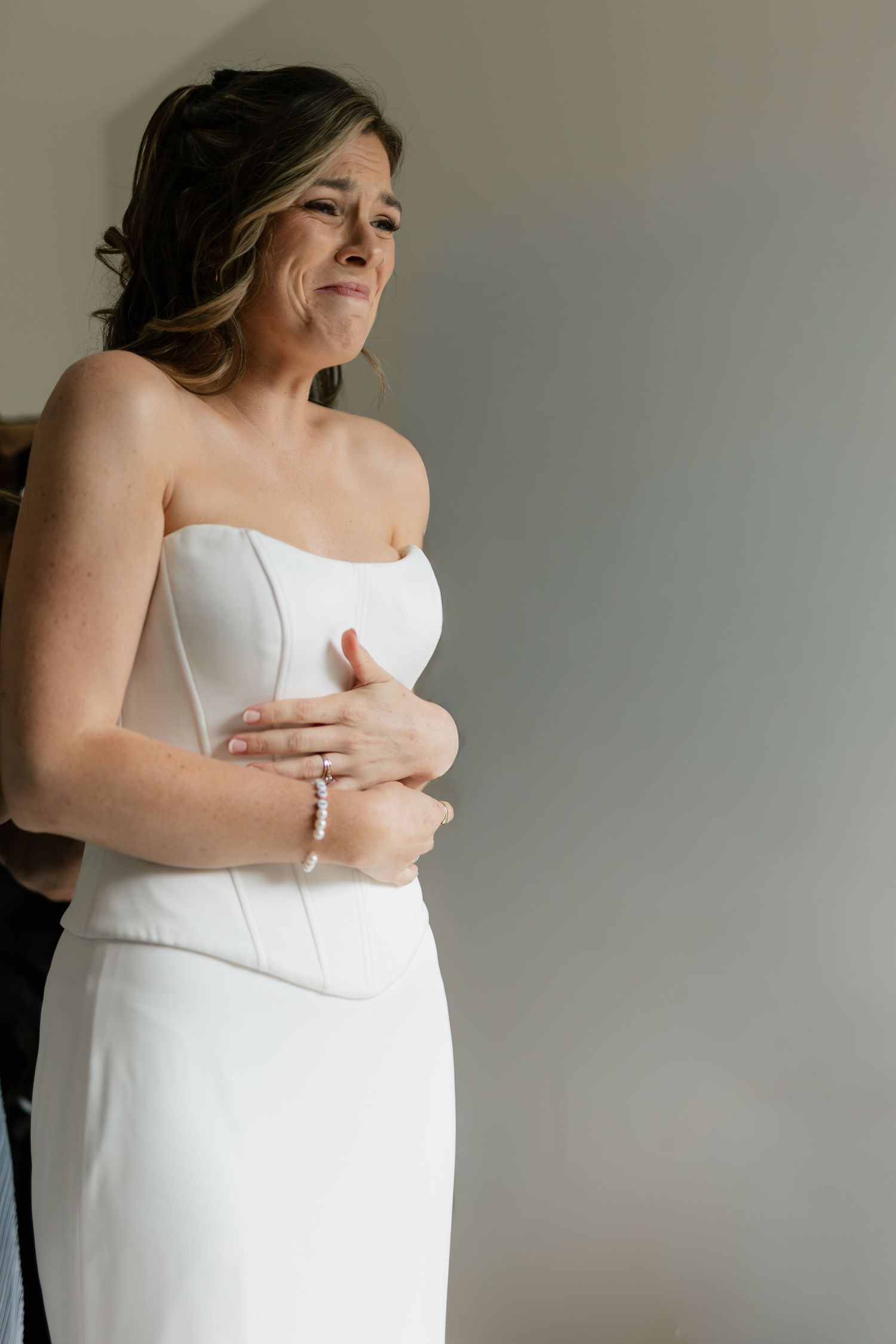 Emotional bride in a strapless white gown being helped into her dress, with tears of joy on her wedding day.