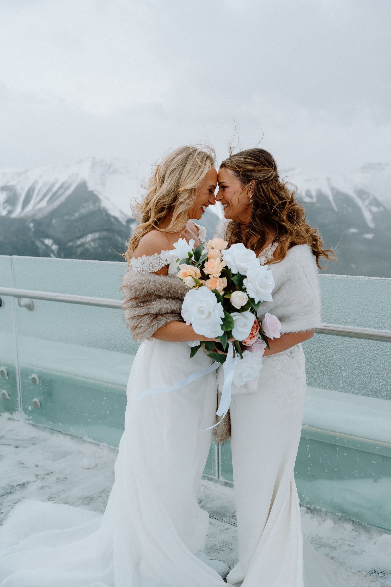 Two brides in white dresses share an intimate moment on a snow-covered mountain overlook.