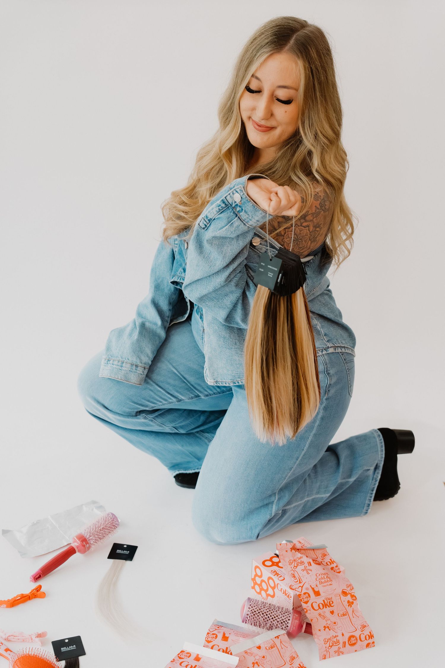 Blonde woman in denim jacket and jeans sitting on floor holding hair extensions with beauty products nearby.