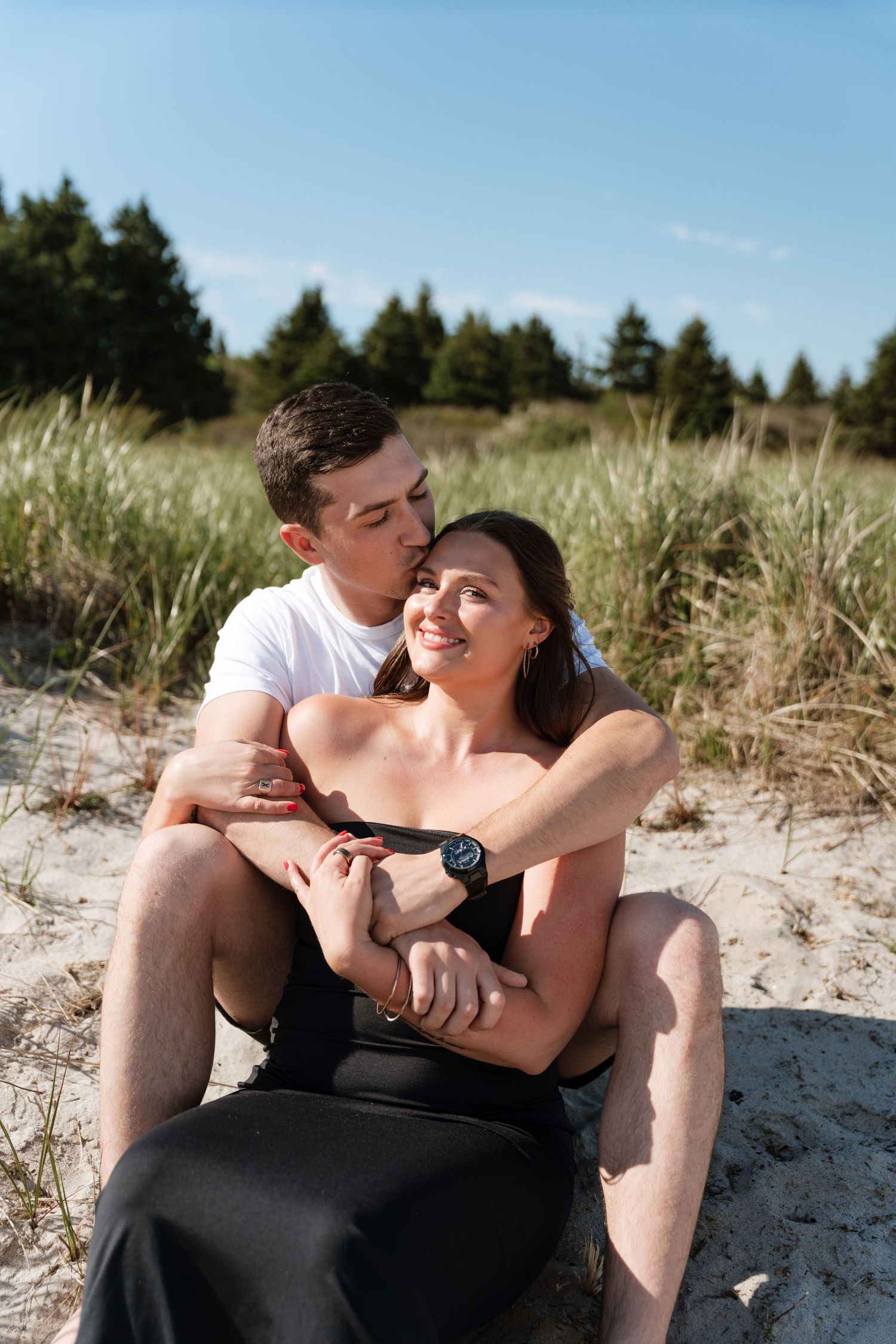 Couple sitting on sandy beach dunes, man kissing woman's cheek, green trees in background on sunny day.