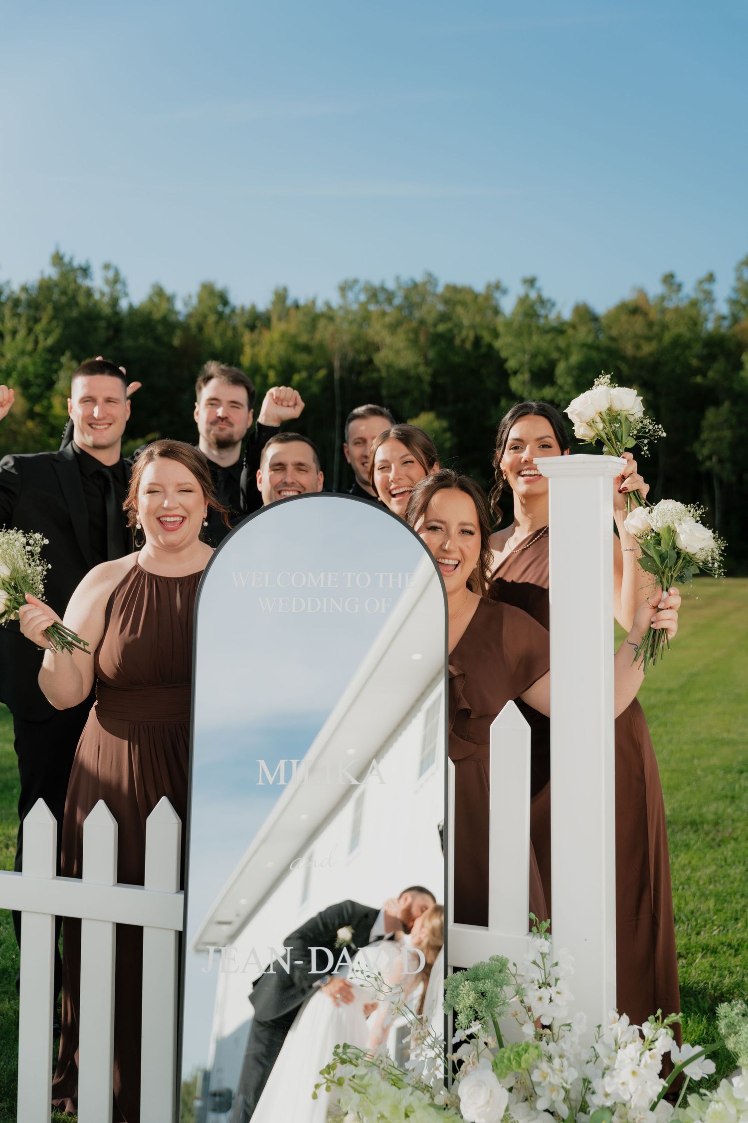 Wedding party in brown dresses posing by white picket fence gate with mirror sign on sunny day.
