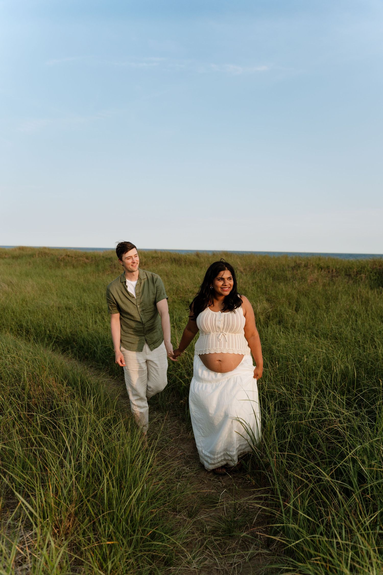 Pregnant couple holding hands walking through green coastal grass field at golden hour, woman in white crop top and skirt.