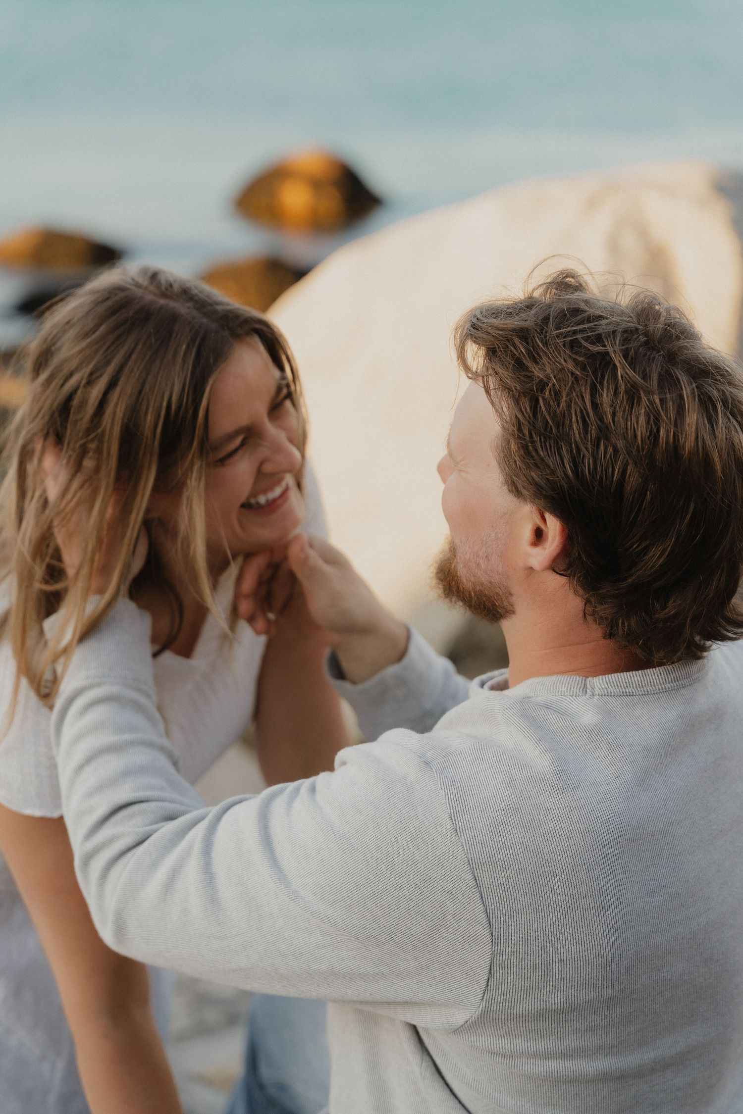 A smiling couple shares a tender moment on a rocky beach, the man gently touching the woman's face.